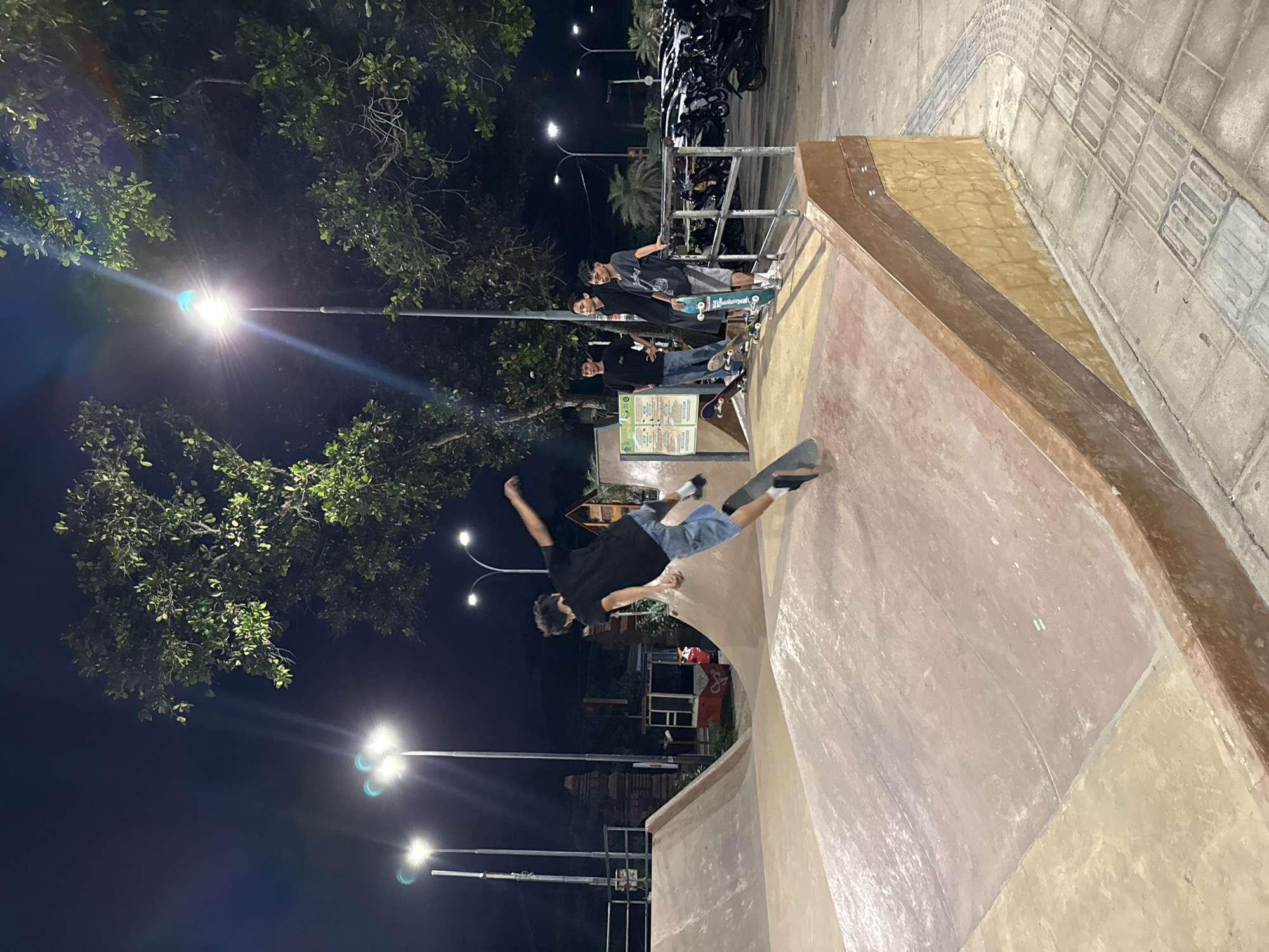 At night in what appears to be a skatepark in Kuta, Indonesia, a person is skateboarding. The individual is wearing a black shirt and light-colored shorts, captured mid-air while performing a trick on a ramp. Other people are present, watching nearby, some with skateboards. The environment includes street lights, trees, a metal railing, a parking area with scooters, and a tiled ground surface.