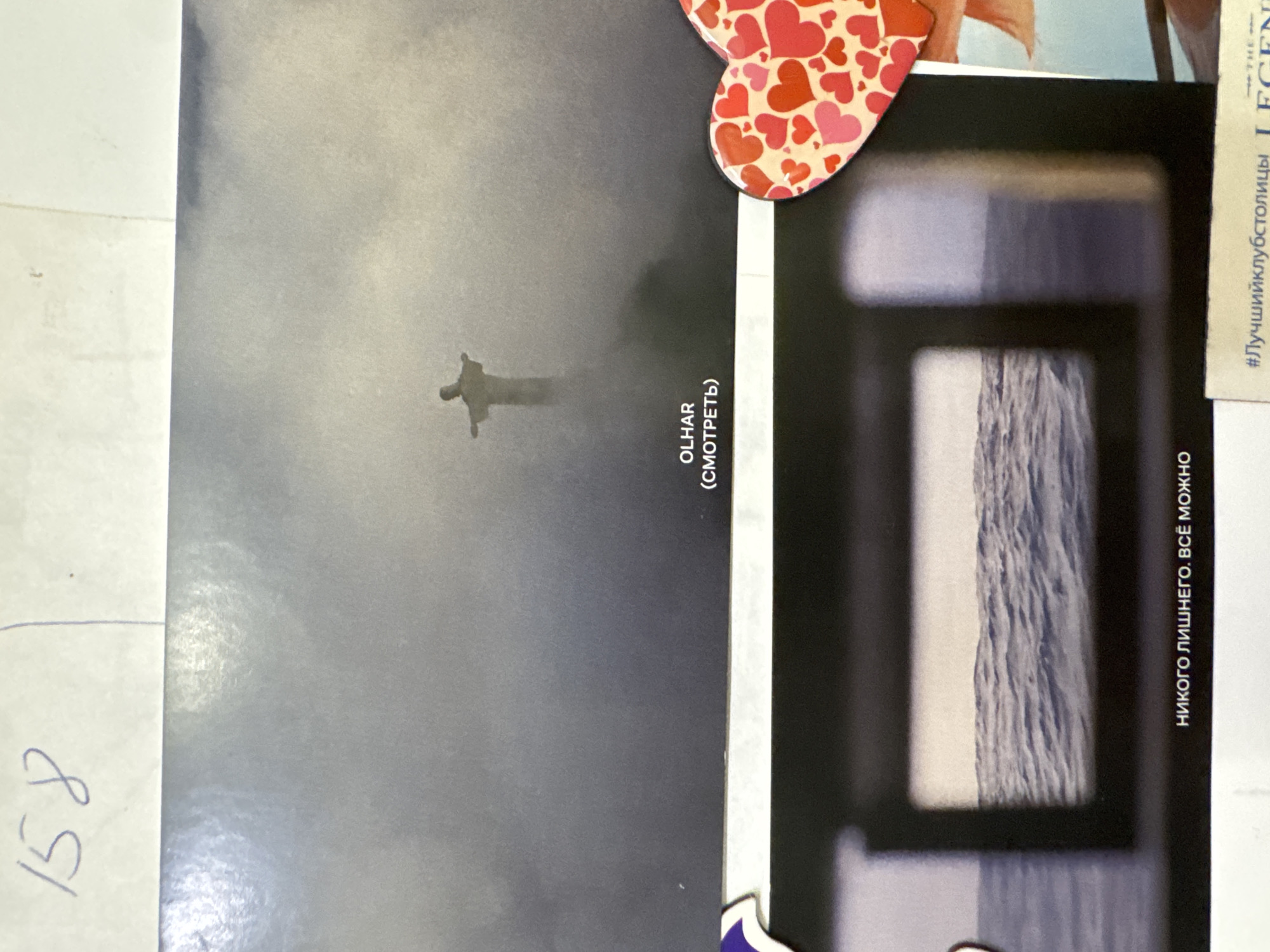 The image shows a collage of photographs. One photo depicts the Christ the Redeemer statue in Rio de Janeiro, Brazil, obscured by fog or clouds. Text adjacent to this image reads "OLHAR (СМОТРЕТЬ)," which translates to "LOOK (WATCH)." Another photo presents a close-up view through a window, possibly showcasing a mountain range or geological formation. A heart-shaped object with red and pink heart patterns is attached to the collage. Text in Russian on the right side reads "НИКОГО ЛИШНЕГО. ВСЁ МОЖНО", meaning "No one extra. Everything is possible." There is a handwritten note "158" on a white background in the bottom left of the image. The scene does not take place in Kazan, Russia.