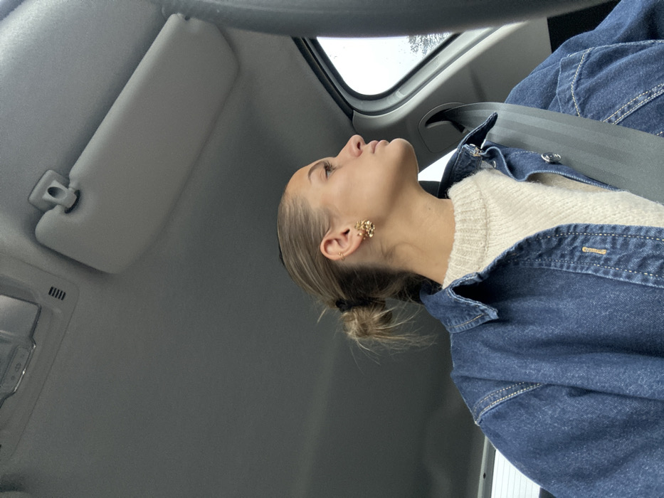 A young adult female is depicted in the front passenger seat of a vehicle, located in Saint-Cyr-sur-le-Rhône, France. She is seen from the chest up, facing right and looking out the side window, with her profile clearly visible. Her light brown hair is pulled back into a messy bun, secured with a black elastic tie. She has multiple piercings on her left ear, featuring a prominent gold-toned, floral or clustered earring on the lobe and several smaller studs along the helix. She wears a cream-colored, ribbed knit sweater beneath an open, dark blue denim button-up shirt or jacket. A dark-colored seatbelt is fastened across her chest and right shoulder. The vehicle's interior includes a light gray headliner, a light gray sun visor, and an overhead console with a light fixture. The exterior environment visible through the window is indistinct and appears overcast or rainy. No other individuals or direct interactions are visible.