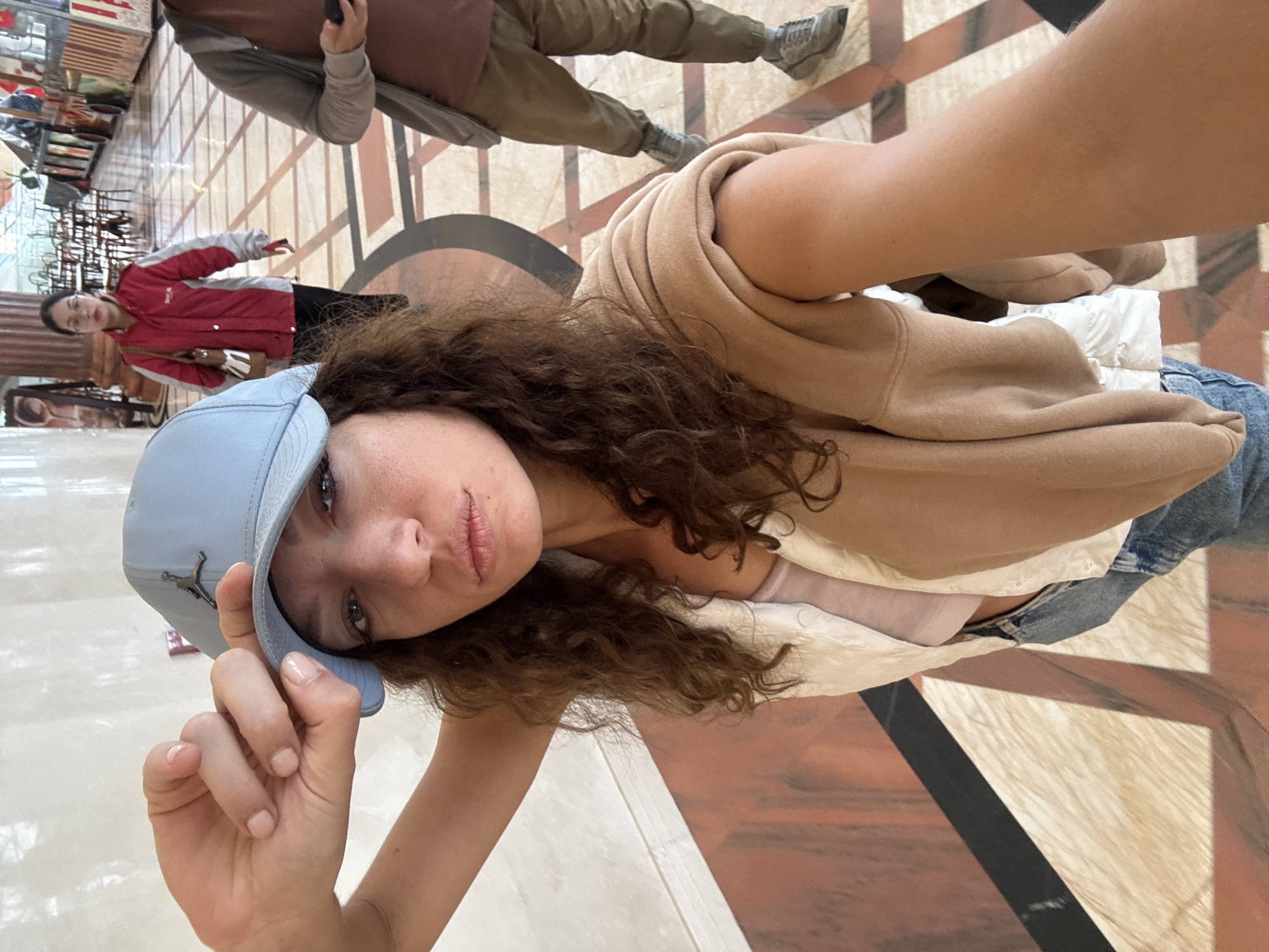 The image captures a selfie or close-up shot of a young woman in an indoor public space, likely a shopping mall in Đà Nẵng, Vietnam. The photo is taken from a low angle, with the subject looking directly at the camera.
**Subjects:** The primary subject is a young woman with long, dark curly hair, wearing a light blue baseball cap with a dark grey Jordan logo. She has light-colored eyes and wears light pink lipstick. She is dressed in a beige long-sleeved top or hoodie and blue jeans. Her fingernails are painted a light nude pink. In the background, two other individuals are visible: a woman in a red and white jacket and dark pants, and a partially seen man in olive green attire and grey sneakers.
**Activity or event taking place:** The main subject is posing for a photograph, possibly a selfie. The background individuals appear to be walking or standing in the public space.
**Notable details:** The setting is characterized by a polished floor with a distinctive geometric pattern of beige, brown, and black tiles, suggesting a modern, well-maintained commercial or public building. Reflections on the floor indicate a glossy surface. The lighting appears to be artificial, consistent with an indoor daytime environment. The main subject has a calm, slightly serious expression.
**Visible text:** The prominent dark grey Jordan logo is visible on the light blue baseball cap worn by the main subject.