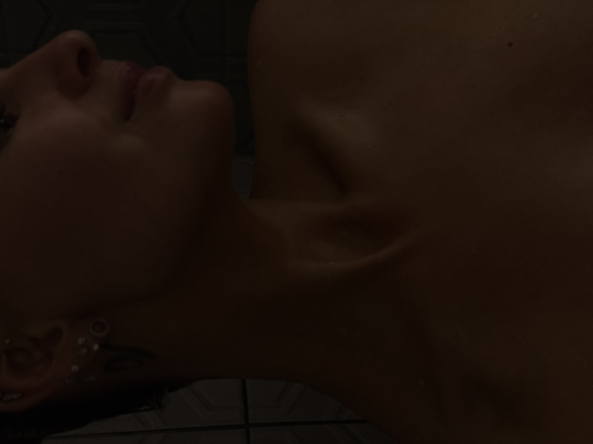 The media file features a close-up portrait of a person, likely female, captured in a dimly lit setting. The subject is shown from the nose and lips down to the upper chest and shoulder, with their head gently tilted back and to the side. The overall color palette is dominated by dark, warm, and muted tones, emphasizing the soft contours of the body.
The person appears to be in a state of rest or relaxation. Their skin has a subtle sheen, suggesting it might be moist from water or sweat. The visible facial features include the profile of the nose and full, slightly parted lips. Part of the right eye is discernible in the upper left corner, looking upwards. A prominent detail is the left ear, which is heavily adorned with multiple piercings, including a large tunnel or gauge in the lobe and several smaller studs along the cartilage. Below the ear, a faint, dark marking, possibly a tattoo, can be seen. The collarbones are clearly defined, indicating a slender build.
The background is largely obscured by shadow, but a faint, dark geometric pattern, suggestive of tiles or a textured wall, is visible behind the person's head and shoulder. The low lighting creates an intimate and subdued atmosphere. No text is visible in the image. The provided location context for this media file is Chaikovsky, Russia.