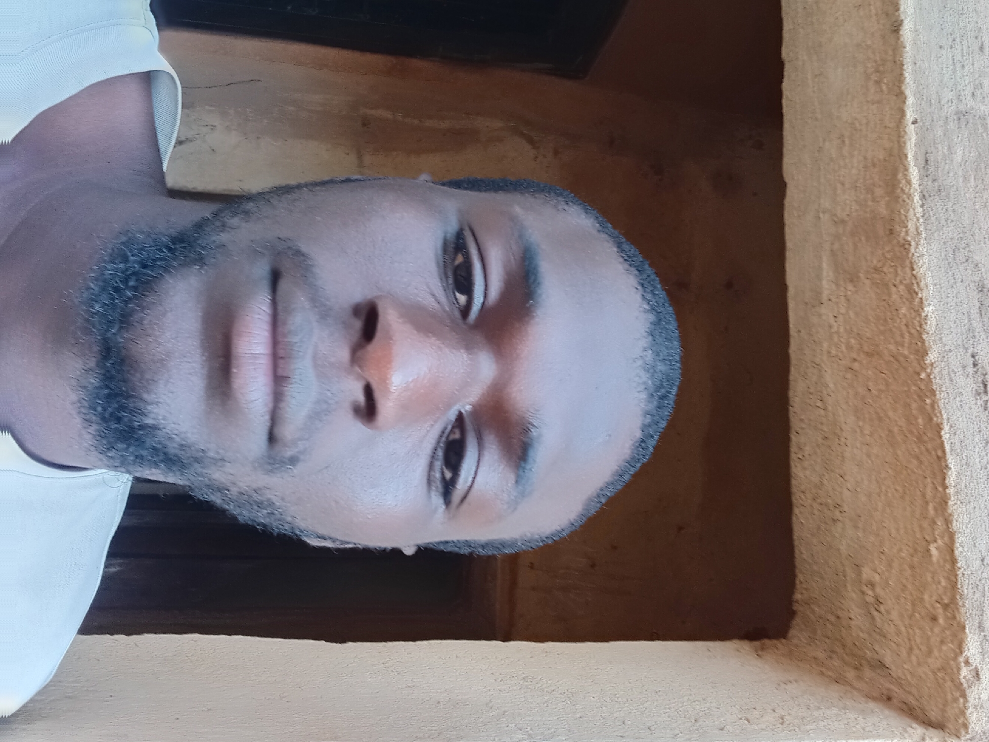 The image is a close-up, front-facing selfie of a young Black man. He has dark skin, short, dark hair, and a light beard and mustache. He is wearing a light-colored, possibly white or cream, t-shirt with a textured neckline. The background appears to be an outdoor environment with rough, textured walls in earthy tones, suggesting an unfinished or rustic building. The lighting is natural and comes from above, casting shadows on his face. The man's eyes are looking directly at the camera. The composition is focused tightly on his face and upper torso. The presence of what appears to be a doorway or opening behind him, framed by the rough walls, suggests he might be standing in an entryway or alcove. Given the prompt's specificity, if this image were taken in Bukuru, Nigeria, the architectural style and material of the walls might be indicative of common building practices in that region.