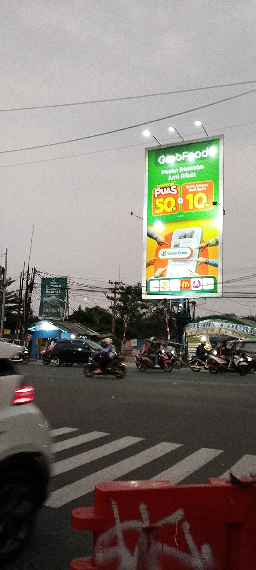 The image depicts a busy street intersection in Depok, Indonesia, likely during the late afternoon or early evening due to the dim lighting. Dominating the scene is a large, vibrant green billboard advertising GrabFood, featuring a promotion of "Diskon Puas 50%" and "Extra Hemat 10". The billboard also displays logos of various food partners, including McDonald's.
In the foreground, a white car is partially visible on the left, its red taillights illuminated, suggesting it is in motion. The road surface shows a pedestrian crossing with white stripes. A bright red traffic barrier, adorned with white graffiti, is positioned on the right side of the road.
The intersection is filled with various vehicles, predominantly motorcycles, with several riders clearly visible. One motorcycle in the center lane has a rider wearing a light-colored helmet and a striped shirt. Another motorcycle on the right has a rider wearing a dark jacket and a helmet. A black car is also present in the intersection.
In the background, several power lines and utility poles crisscross the sky, indicating an urban environment. To the left of the main billboard, another, smaller billboard with text and an image of a mosque is partially visible. Further in the background, a blue-roofed structure with bright lighting, possibly a shop or stall, is visible, with people gathered around it. Trees and foliage are scattered throughout the background, suggesting the presence of green spaces within the city. The overall atmosphere is one of active urban life and commerce.