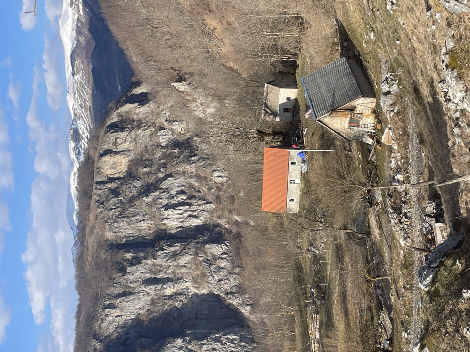 The image depicts a rural mountainous landscape in Mokro, Montenegro, under a clear blue sky with scattered white clouds. Distant snow-capped peaks rise above an extensive, steep grey rocky cliff face and densely forested hillsides primarily composed of bare deciduous trees. In the mid-ground, nestled on a sloping, partially cleared terrain, are three stone-built structures.
The most prominent building, positioned centrally, is a two-story stone house with a bright orange-red gabled roof, white-framed windows, and a white front door. To its right, two individuals are engaged in activity; one is crouched near a blue tarp on the ground, while another stands nearby. A vertical pole, possibly a utility or flag pole, stands in front of this house. Behind this structure, partially obscured by trees, is a smaller, older stone building with a darker, gabled roof.
In the lower right foreground, a third rustic stone building features a corrugated grey metal gabled roof and a wooden upper facade. A wooden ladder leans against its front. The immediate foreground consists of dry grass, scattered rocks, small bare trees, and visible fencing constructed from wooden posts and wire to the left of the central house. The ground around the structures exhibits signs of disarray, including scattered rocks, debris, and what appears to be stacked materials.