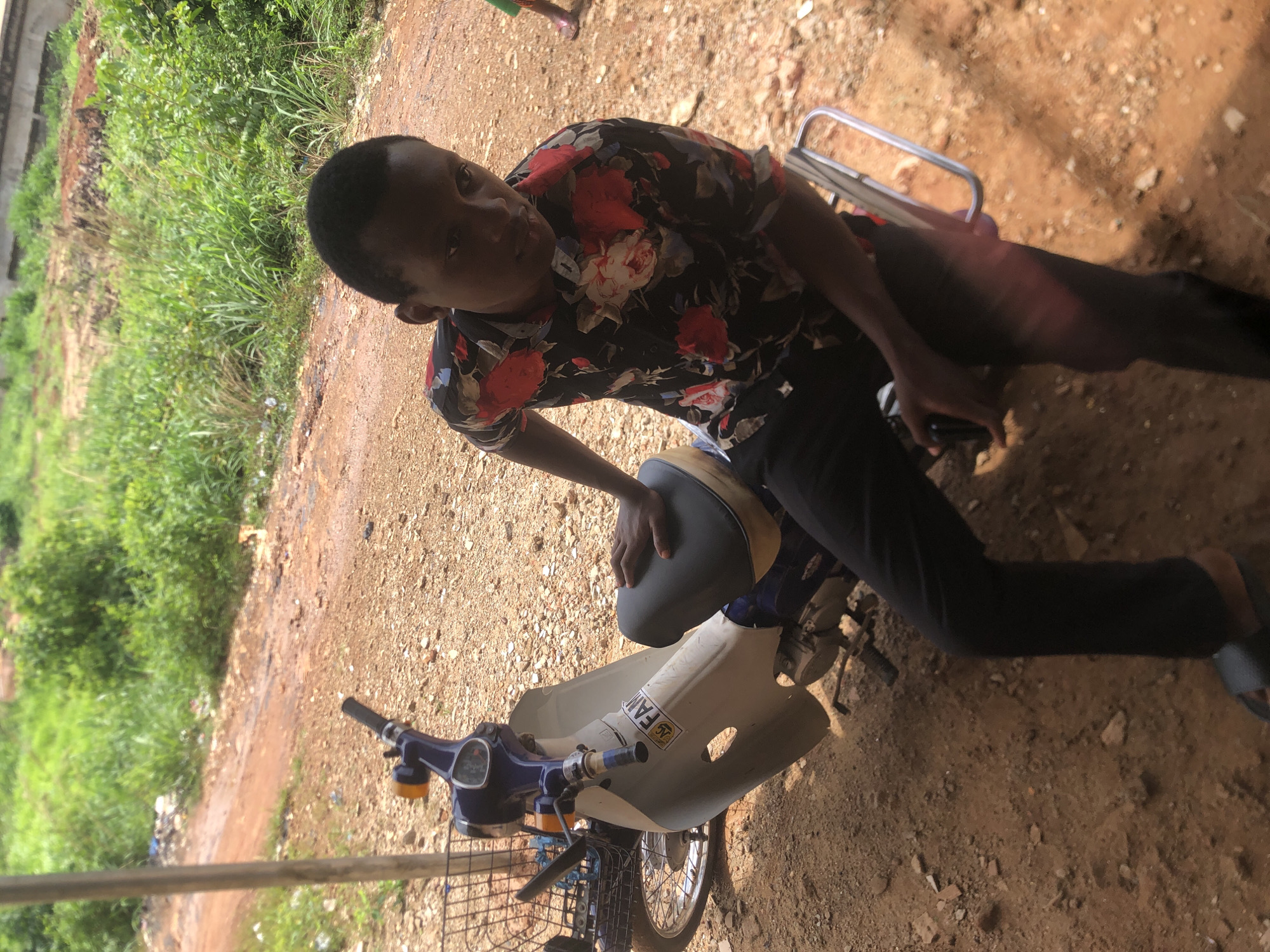 A young man in a floral shirt and dark pants is sitting on a light-colored motorbike with a front basket in an outdoor setting in Nkwelle, Nigeria. The ground is dirt. Vegetation and a concrete structure are visible in the background. The man is looking at the camera.