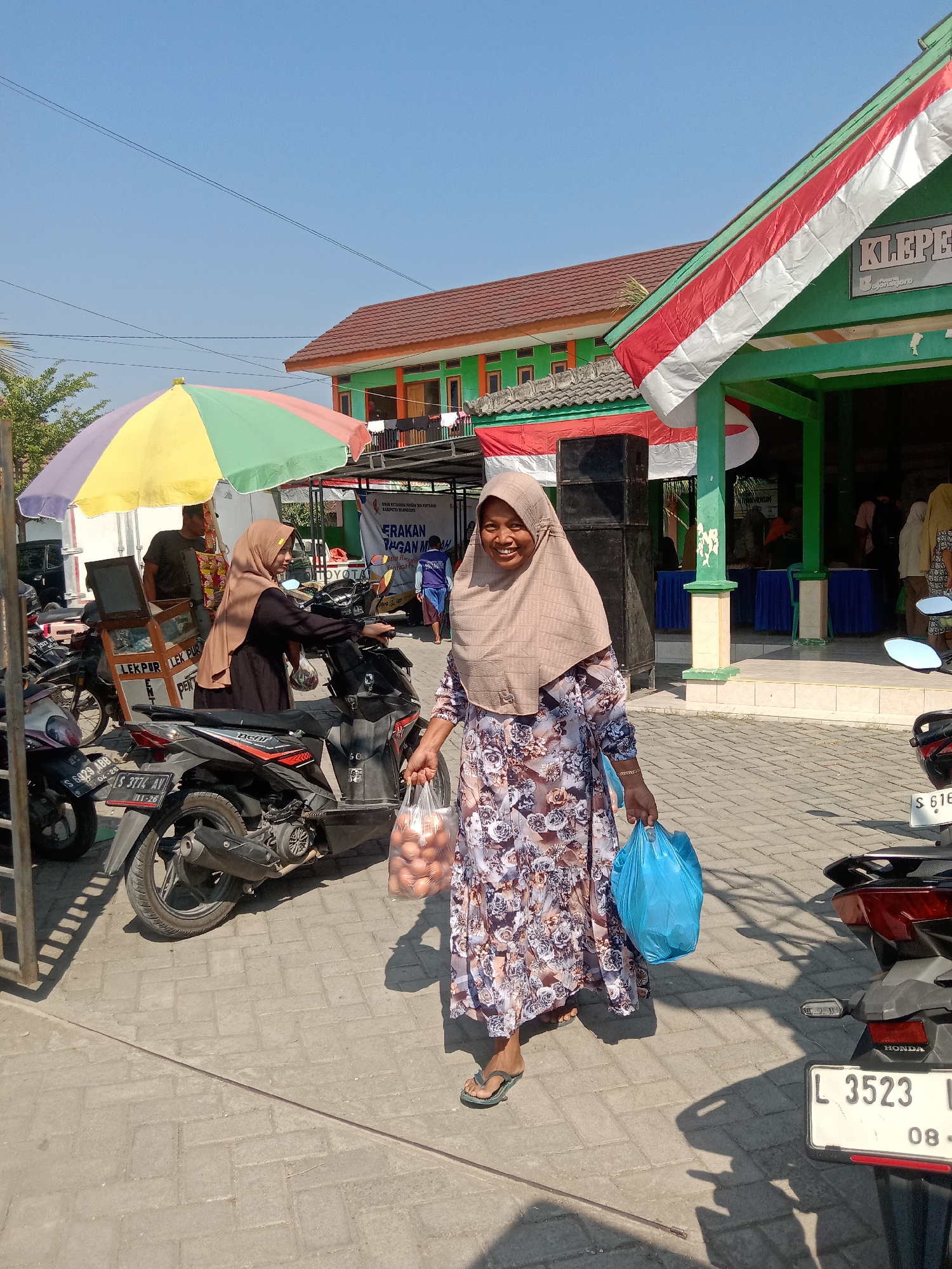 A woman wearing a light brown hijab and a long, floral-patterned dress is walking towards the camera, smiling. She is carrying a clear plastic bag filled with eggs in her right hand and a bright blue plastic bag in her left hand. She is wearing light green flip-flops. In the background, a green building with red and white Indonesian flags draped over its entrance is visible. The building has the word "KLEPE" visible on a sign. Several motorcycles are parked on a paved area in front of the building. A colorful umbrella is set up to the left of the woman, and a man is partially visible behind it. The sky is clear and blue. The scene appears to be outdoors in a public area, possibly a market or community center, in Klepek, Indonesia.