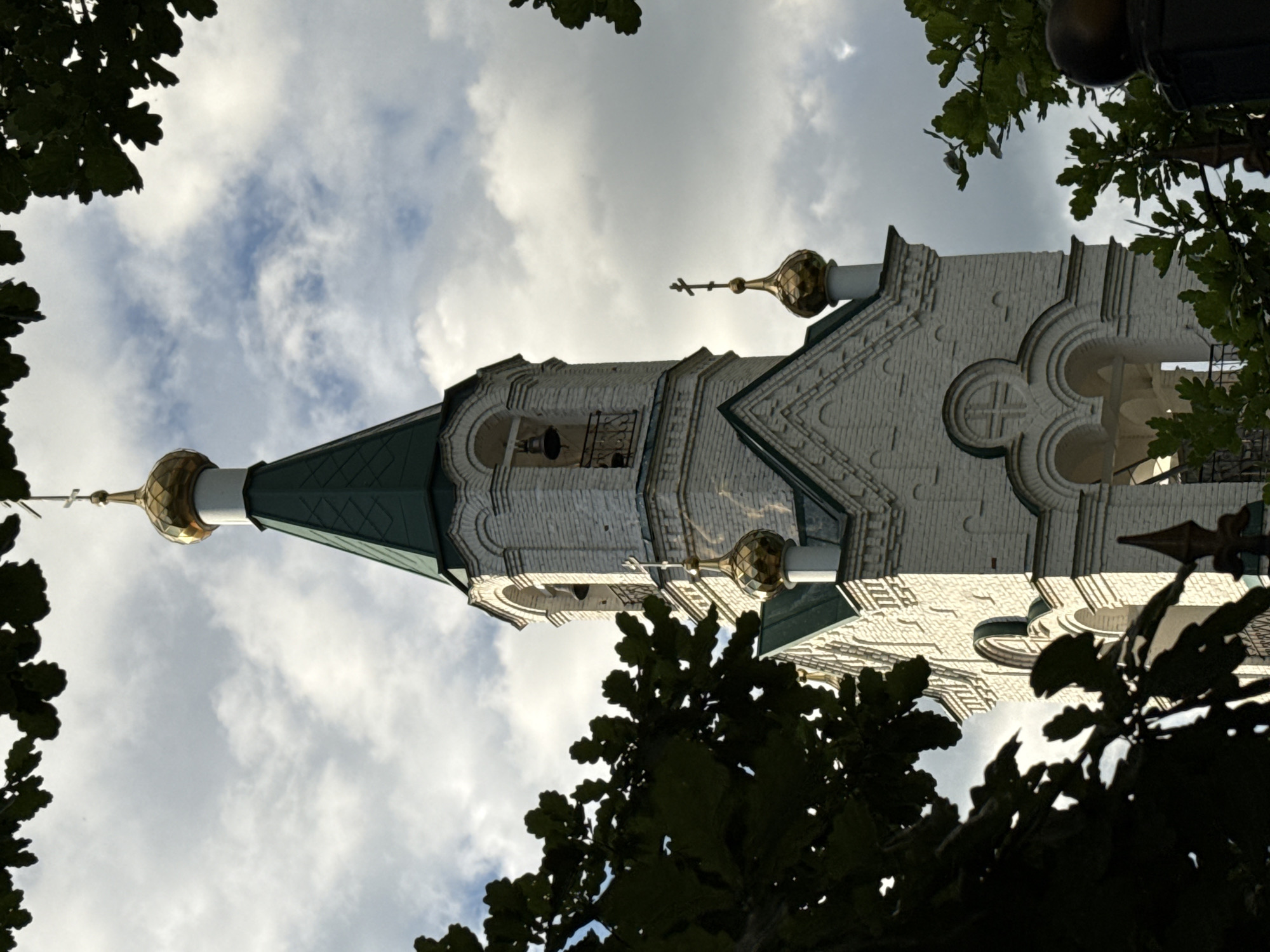 The image shows a low-angle shot looking up at the bell tower of a white brick church, framed by dark green foliage. The church has a green, conical roof topped with a golden dome and cross. There are also smaller golden domes on the structure, some with crosses. The architecture features decorative white brickwork with arched windows and patterned designs. A bell is visible through one of the windows. The sky is partly cloudy, with bright patches suggesting sunlight breaking through. The time of day appears to be late afternoon or early evening, given the quality of light. The overall mood is serene and perhaps a little solemn. The image is a close-up view of the church tower, with the trees providing a natural frame and adding a sense of depth. No people or text are visible in the image. The context of Votkinsk, Russia, suggests this is a religious building within that city.