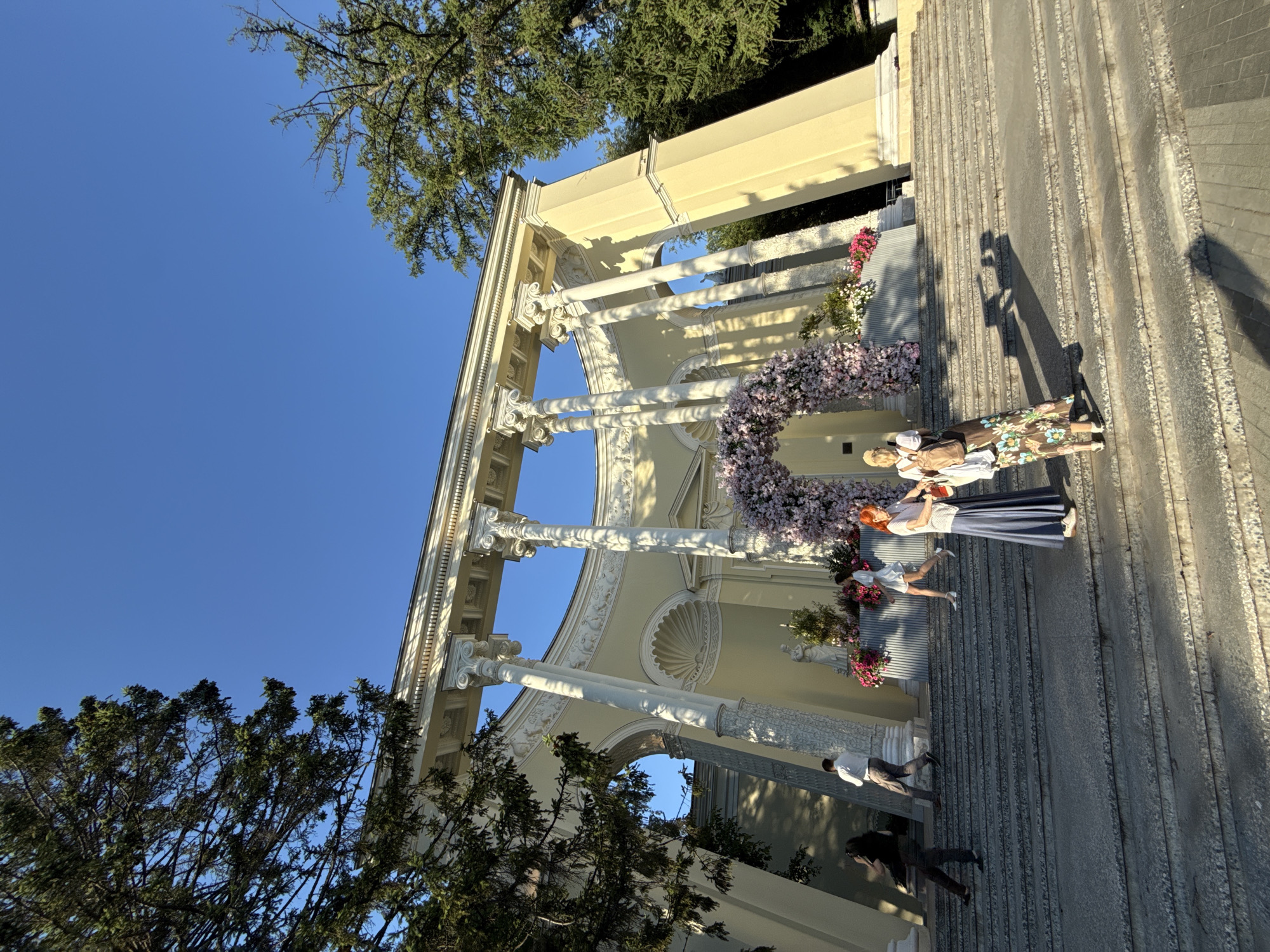 The image shows the colonnade of the National Agricultural Exhibition Center in Moscow, Russia. The structure is neoclassical, with large white columns supporting a decorative roof. In front of the colonnade, there is a large floral archway adorned with pink and white flowers. Several people are visible on the steps leading up to the colonnade. Two women are standing in the foreground, facing each other and engaged in conversation. One woman has bright red hair and is wearing a white shirt and a long blue skirt. The other woman is wearing a white top and a floral dress. Behind them, a woman in a white dress is walking up the steps. Further up the steps, a man in a white shirt and dark pants is also walking. In the background, a statue is visible behind the floral arch. The scene is illuminated by sunlight, casting shadows on the steps. The sky is clear and blue.