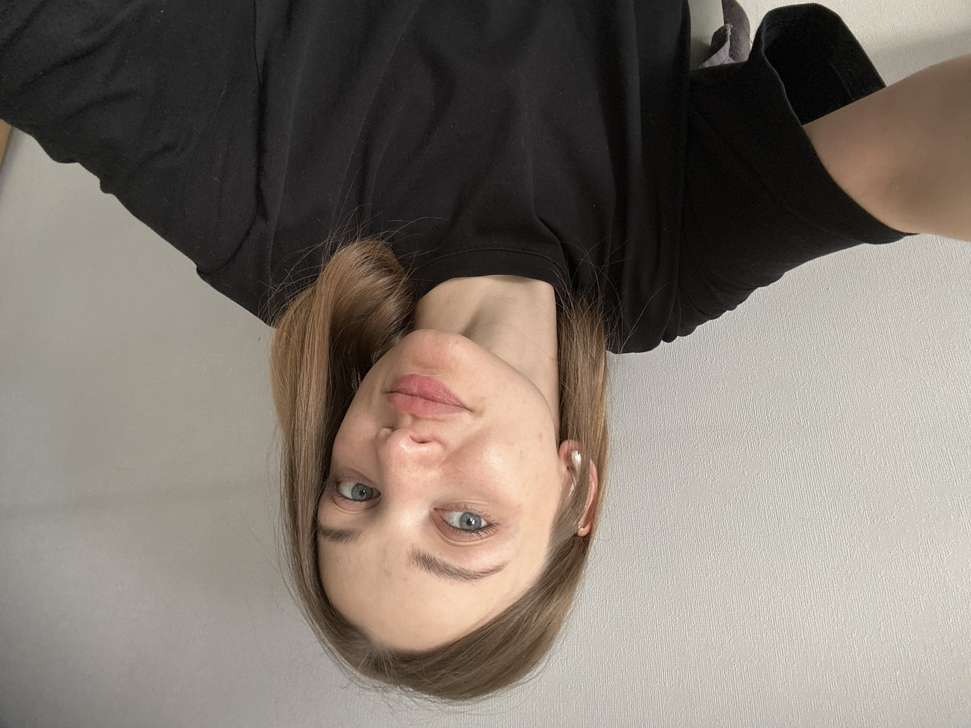 The media file displays an inverted (upside-down) portrait of a young woman. Her light brown hair, parted on the side, falls around her face. She has light blue or grey eyes, which look directly at the camera with a neutral, slightly pensive expression. Her lips are naturally pink and softly closed. She appears to have clear skin, with some minor redness visible on her forehead and cheeks.
She is wearing a plain black crew neck t-shirt with short sleeves. A white wireless earbud is visible in her right ear. The background is a simple, light grey wall, which exhibits a subtle vertical texture, suggesting wallpaper. The lighting is bright and even, indicating an indoor setting, possibly during the day. No explicit location cues are visible within the image itself to confirm the provided context of Kazan, Russia. There is no visible text in the image.