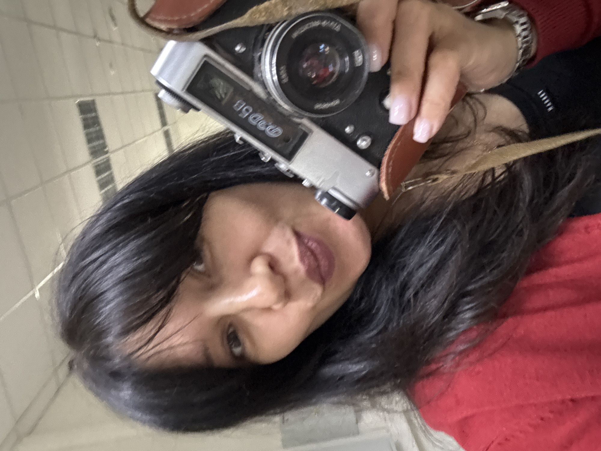 An indoor, vertically oriented close-up selfie captures a woman holding a vintage camera. The woman, with long dark hair, gazes towards the viewer with a neutral expression, her lips painted a dark red. She wears a red sweater over a black garment, which has white text "URBAN" visible. A metallic watch adorns her wrist. Her hand, featuring light pink manicured nails, holds a classic silver and black "FED 50" camera with a brown leather strap. Part of the camera lens shows text including "INDUST**" and "4/50". The background is a light-colored tiled or paneled wall, likely illuminated by indoor lighting. The image suggests a personal moment, possibly taken in Chaikovsky, Russia, highlighting the retro camera.