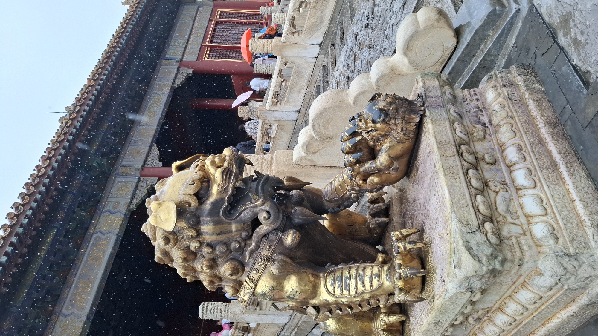A close-up, low-angle shot depicts a golden bronze lion statue in front of a building in Dongcheng District, China. The lion has an ornate, patterned mane and body armor. The building behind it has a dark roof with upturned eaves decorated with multiple levels of tiles. There are red support beams and window frames on the second level of the building. People holding umbrellas can be seen behind a stone balustrade of carved, rounded pillars. Snow is falling. The lion is positioned on a heavily carved stone pedestal.