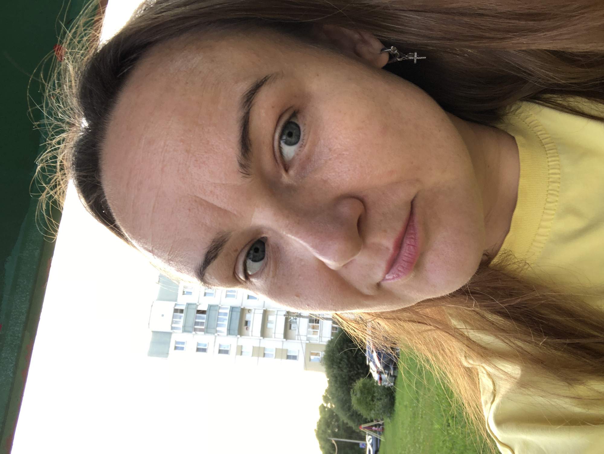 A woman with light brown hair and blue eyes takes a selfie outdoors in front of a residential building. She is wearing a yellow t-shirt and a silver cross earring. The background shows a tall, multi-story apartment building with a green balcony on the fifth floor, a grassy area with trees, bushes, and parked cars. A playground structure is visible in the distance. The sky is bright and overcast. The scene appears to be in the city of Rusanovo, Russia.