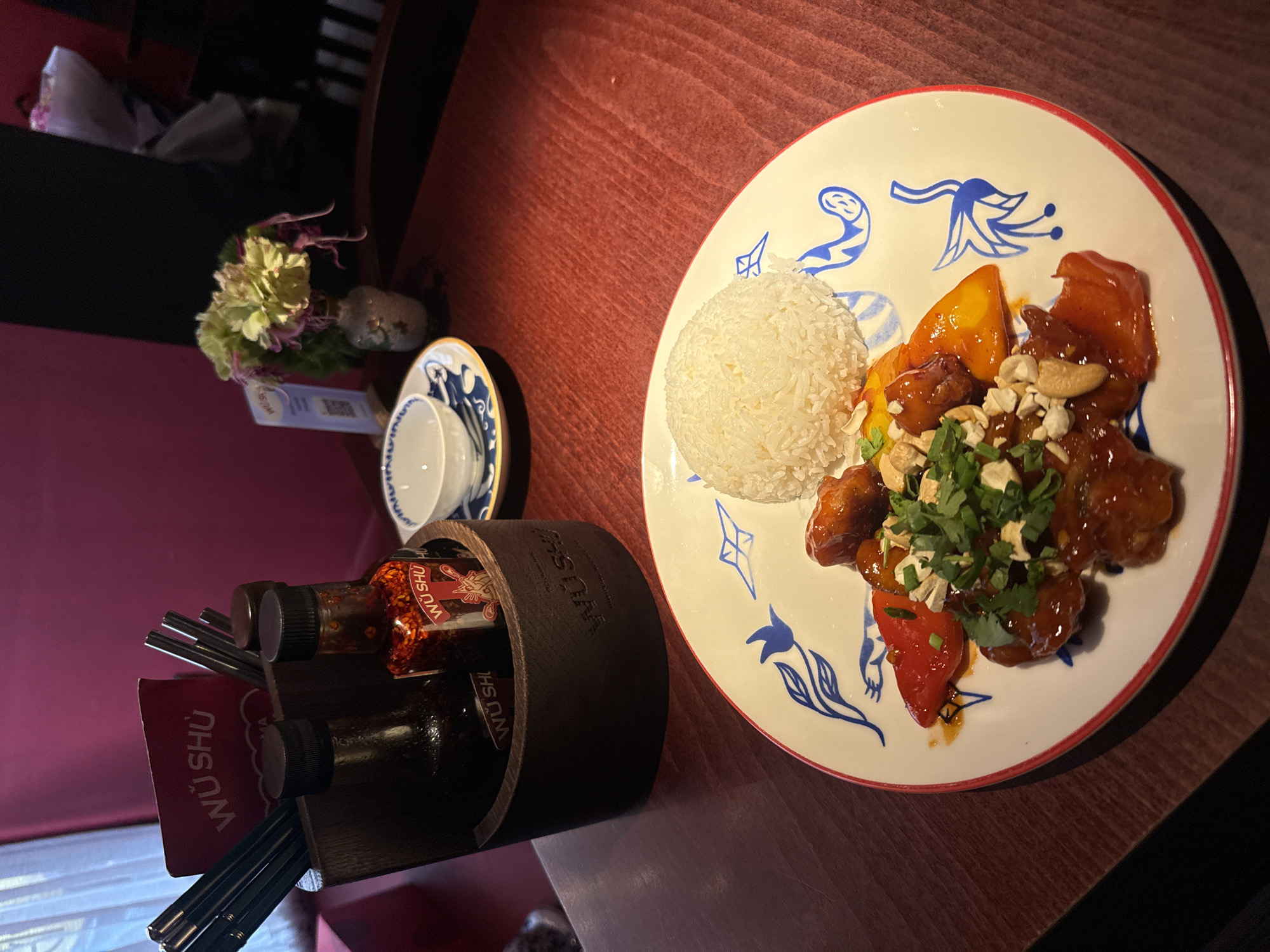 The image captures a meal served in an indoor restaurant setting, likely in Moscow, Russia. The central focus is a white plate adorned with blue decorative patterns and a red rim, holding a portion of food. On the plate is a mound of white rice and a dish consisting of golden-brown glazed pieces of what appears to be chicken or a similar protein, mixed with red and yellow bell peppers. This dish is generously topped with chopped cashews and fresh green herbs, likely cilantro.
To the left of the plate, a small, white teacup with blue patterns sits on a matching saucer. Further left, a wooden caddy holds several pairs of black chopsticks and two small bottles of sauce. Visible text on the sauce bottles and a red card within the caddy reads "WÚ SHÙ", which is likely the brand name of the restaurant or condiments.
In the background, a small floral arrangement with light green and purple flowers in a tiny white vase adds to the decor. The table surface is a reddish-brown wood. The lighting appears to be artificial, suggesting it is evening or late afternoon. No people are visible in the image.