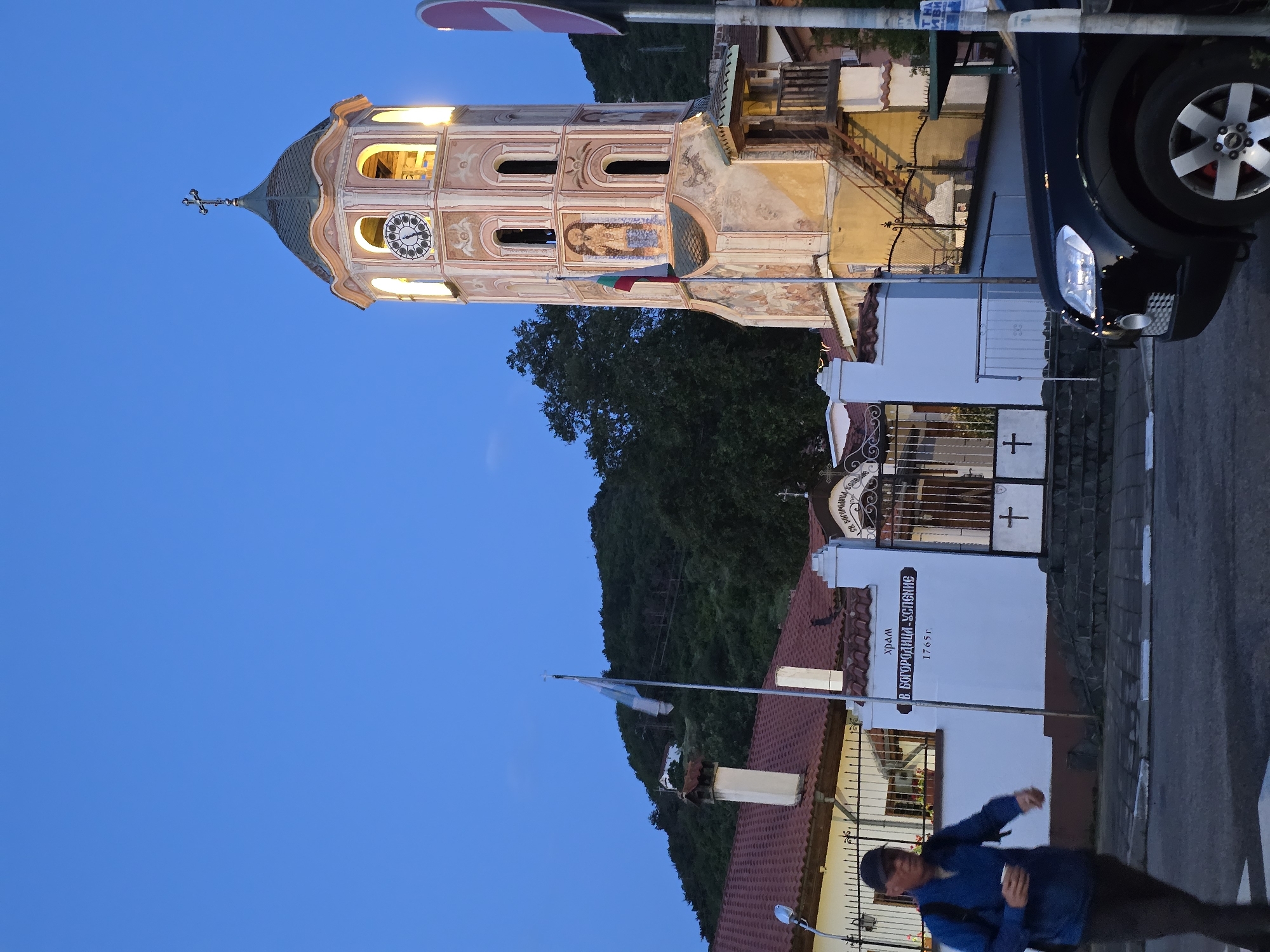 The image, currently rotated 90 degrees clockwise, captures an evening scene featuring an illuminated Orthodox Christian church bell tower. The sky is a deep blue, indicating dusk or nightfall.
The central subject is a tall, ornate bell tower with a domed roof topped by a cross. The tower, constructed from light-colored stone, is adorned with reddish-brown decorative patterns and features arched windows brightly lit from within. A clock face is visible on one side, and religious frescoes, including one depicting a figure below the clock, decorate its exterior. A Bulgarian flag, with horizontal green, white, and red stripes, hangs from the tower.
To the right, a dark-colored car, likely an SUV or van, is partially visible on a paved street, with its front headlight illuminated. In the foreground, a person, appearing to be male and wearing a blue jacket and a dark cap, walks on the pavement, holding a phone.
Below the bell tower, white-walled buildings with red-tiled roofs form part of the church complex. A prominent white wall features Cyrillic text: "ХРАМ СВ. БОГОРОДИЦА УСПЕНИЕ 1765г.", translating to "CHURCH OF THE HOLY VIRGIN ASSUMPTION 1765". A decorative metal gate with crosses and additional Cyrillic text, possibly "СВ. БОГОРОДИЦА", stands in front of this building. Dark green, tree-covered hills rise in the background under the clear, deep blue sky. The scene suggests a historic and peaceful setting, likely in Bulgaria, due to the visible flag and Cyrillic inscriptions.