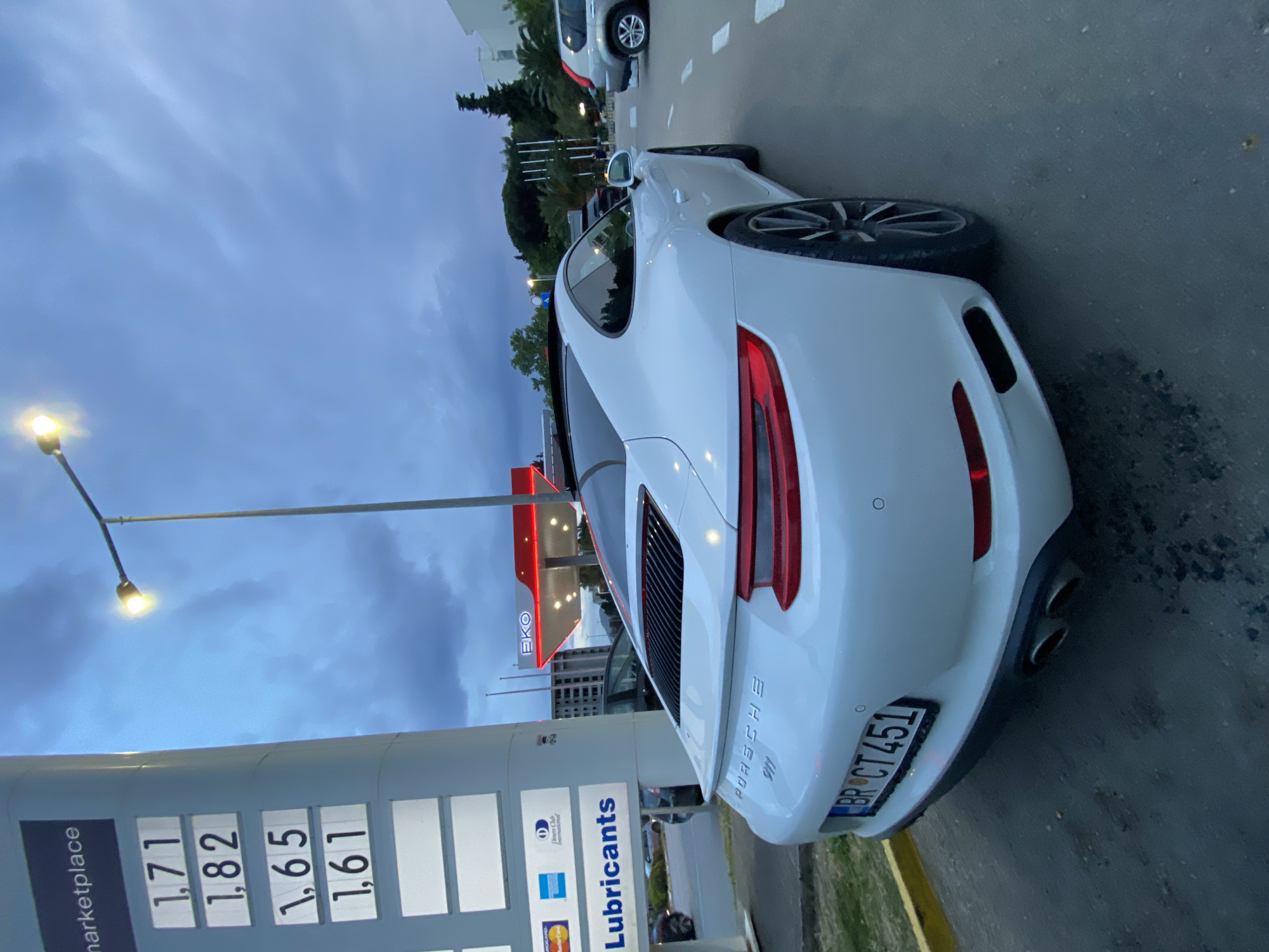 A white Porsche 911 is parked at an EKO gas station in Bar, Montenegro. The gas station's fuel prices are displayed: 1.71, 1.82, 1.65, and 1.61. Various credit card logos and "Lubricants" are visible below the price display. The car's license plate reads "BR CT451". The sky is overcast. Other cars and greenery are in the background.
