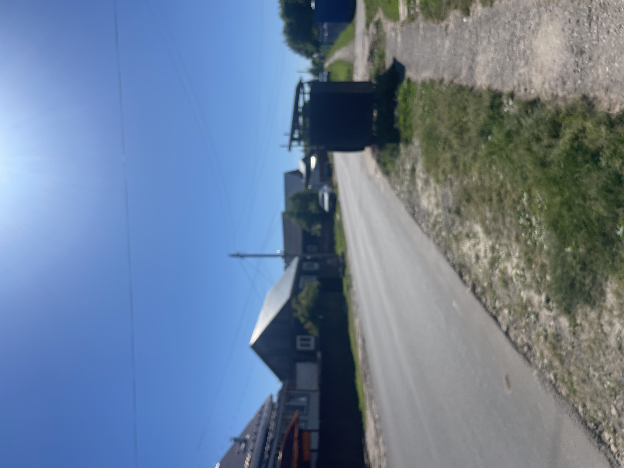 The image depicts a sunny day in a rural area, possibly a village in the Krasnye Baki, Russia. A paved road runs diagonally from the bottom left towards the top right of the frame. On the left side of the road, several houses with dark roofs are visible behind a dark fence. One house in the foreground has a gabled roof and a white-framed window. On the right side of the road, a gravel path runs parallel to it, bordered by grass and weeds. In the foreground on the right, a dark, rectangular structure with a sloped roof, possibly a small shed or utility box, stands partially obscured by vegetation. Further down the road, more houses and trees are visible, along with a white car parked near a house. The sky is a clear, bright blue with some lens flare from the sun. The overall scene is one of a quiet, residential street on a summer day.
