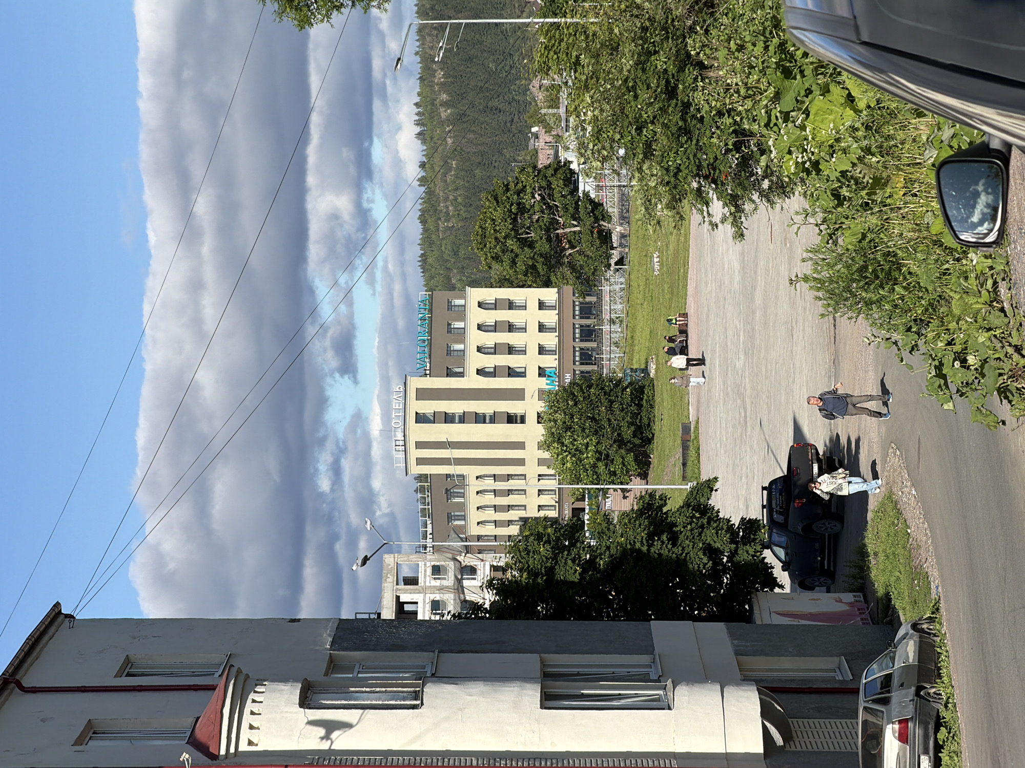 The image captures a view of a street in Sortavala, Russia, with a focus on a building that appears to be a hotel. In the foreground, a grey car is parked on the left, partially obscured by the right side of the frame. A dark-colored pickup truck is parked behind it. Two people are walking on the road in the middle ground, a woman in a light-colored jacket and jeans, and a man in a dark t-shirt and grey pants. Further back, several individuals are seated on benches in a grassy area. Dominating the background is a modern hotel building with a sign that reads "ОТЕЛЬ VALORANTA". The hotel is light yellow with dark brown accents and has multiple floors with numerous windows. Behind the hotel, a dense forest covers a hillside under a sky with scattered clouds. The overall scene suggests a daytime urban environment with elements of residential buildings, businesses, and natural scenery.