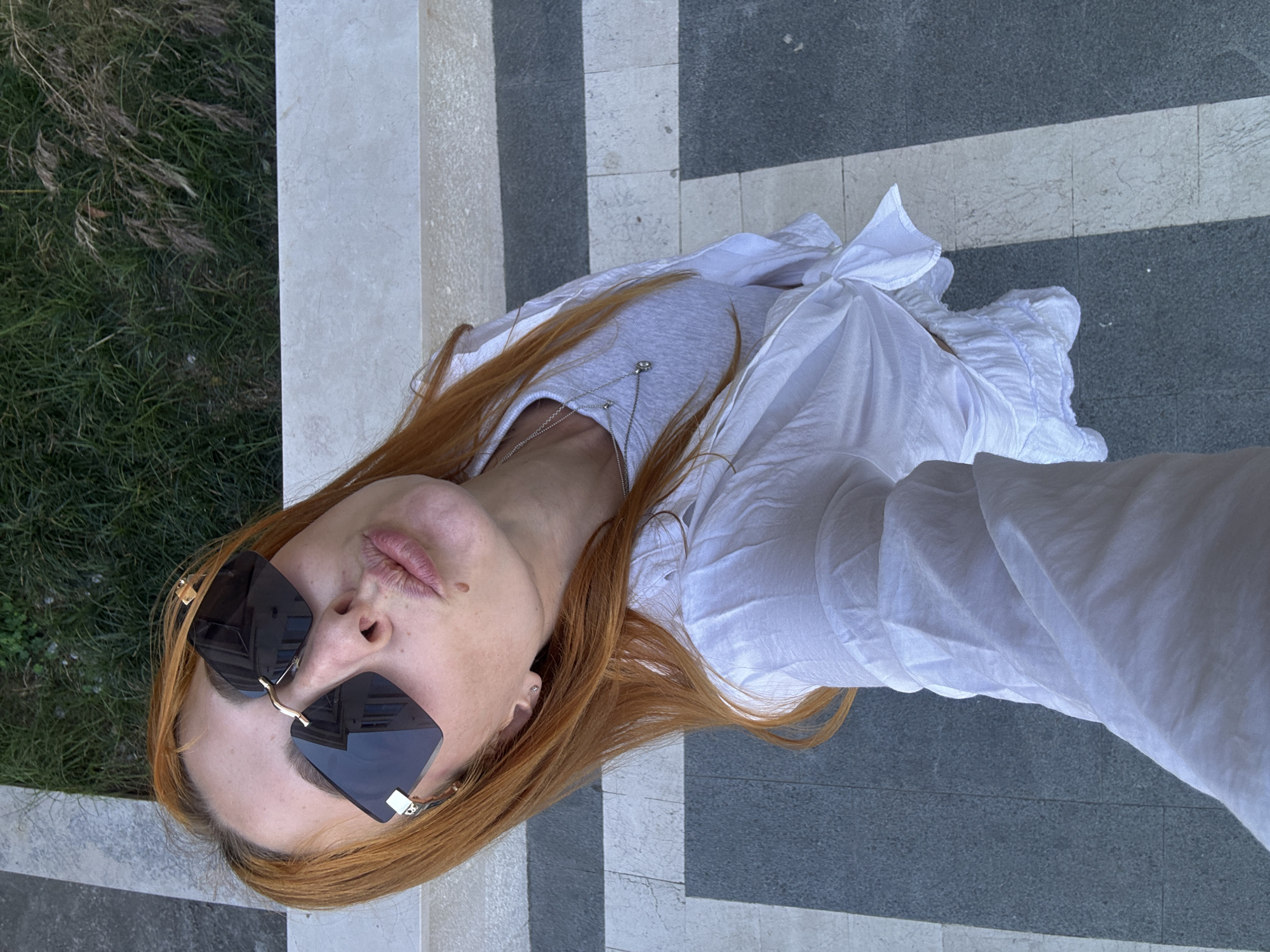 A woman with auburn hair is featured in a high-angle selfie, wearing dark, rectangular sunglasses with metallic frames. She is making a puckering lip gesture. A small mole is visible below her lower lip. She wears a white t-shirt beneath a white, lightweight, long-sleeved top that is tied at the front and features a ruffled lower hem. A silver-toned layered necklace with two small pendants is visible around her neck. The background shows a paved area with alternating light grey and dark grey rectangular tiles. Beyond the pavement, a light-colored stone wall borders an area of green grass mixed with dry vegetation. The scene is situated in Budva, Montenegro.