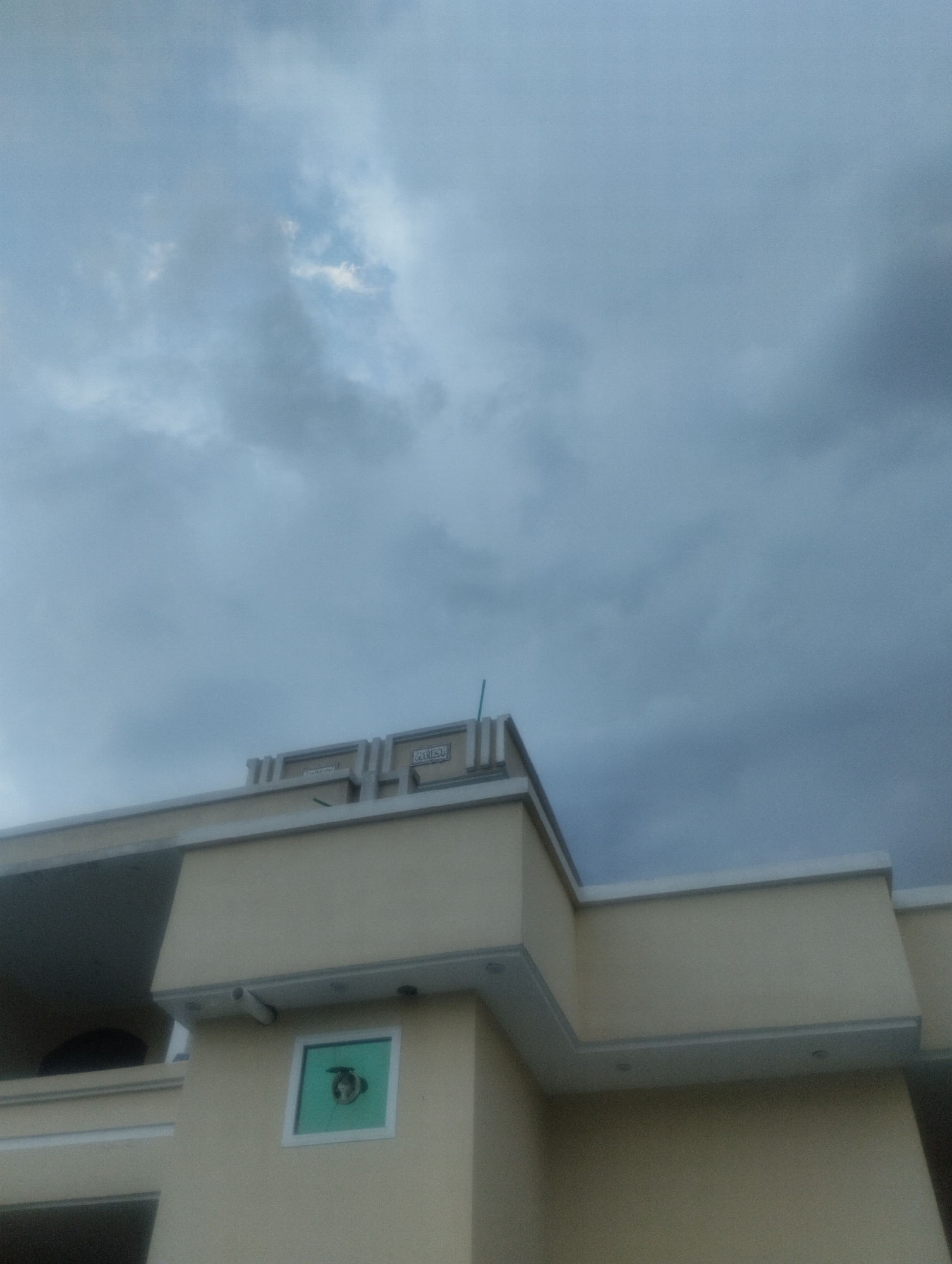 A light tan building with white trim is visible against a cloudy sky. The building has multiple levels, with a balcony area visible on the left side. A small, square, green-tinted window with white trim is present on the middle level of the building. Above the window, a white pipe protrudes from the wall. Decorative elements with embossed text are visible on the upper edge of the building. A thin green pole is also visible extending upwards from the roofline on the right side of the image.