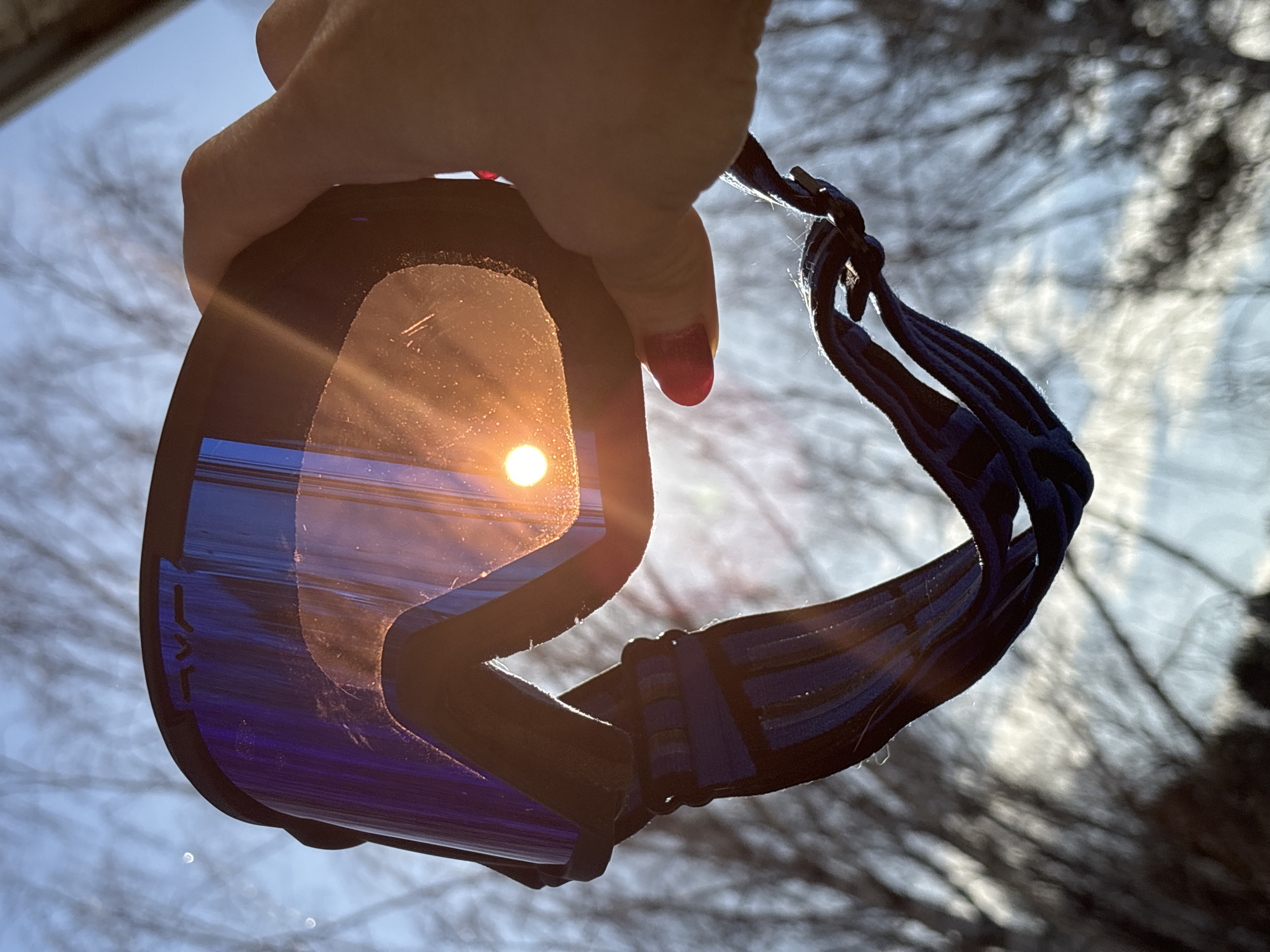 A human right hand, with fingernails painted red, holds a pair of black-framed ski goggles with blue mirrored lenses. The brand "RVE" is visible on the upper frame of the goggles. A blue and black patterned fabric strap hangs vertically from the goggles. The sun is positioned behind the goggles, generating a distinct orange circular light visible through the right lens and creating a prominent lens flare. The background consists of a clear, light blue sky and out-of-focus bare tree branches, indicating an outdoor, leafless environment. This scene is located in Chaikovsky, Russia.