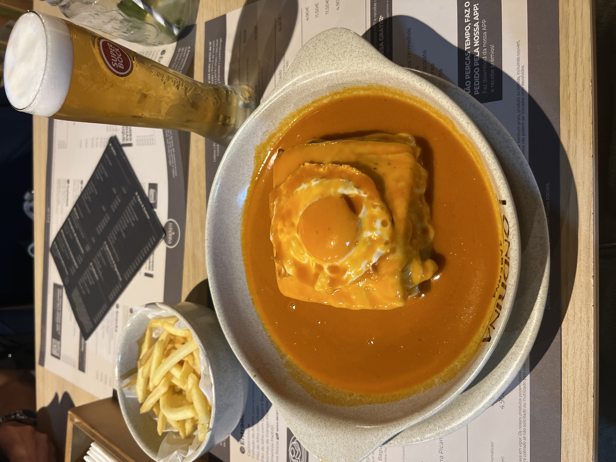 A close-up, eye-level shot captures a French-Portuguese dish, likely a Francesinha, in a pale grey, shallow bowl. The dish is covered in a rich, orange-red sauce and topped with a fried egg with a runny yolk. Alongside the main dish, a small, grey bowl contains a portion of golden french fries. In the background, a tall glass of amber-colored beer with a frothy head sits on a wooden table, next to a menu. The environment suggests a restaurant setting, with hints of a menu and wooden table surfaces. The focus is on the food and drink, presented in a visually appealing manner.