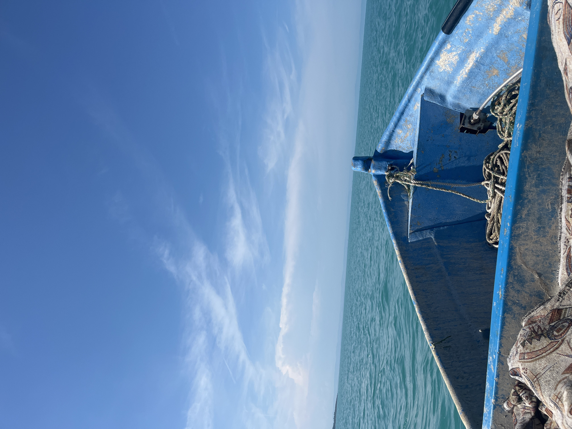 The image presents a forward-facing view from inside the bow of a worn, blue utility boat on a body of water at an undisclosed location. The boat's interior, visible in the lower half of the frame, displays faded and chipped blue paint, revealing lighter patches. Coils of white rope are stored in the front compartment of the boat, near a metal fitting at the bow. A patterned textile or blanket is partially visible in the bottom-left foreground, covering part of the boat's interior. A metallic tube or hose runs from the right side of the boat into the central compartment.
The water, occupying the middle section of the image, is a clear turquoise color, exhibiting small surface ripples that indicate calm to slightly choppy conditions. It extends to a distant, hazy horizon line. A low-lying, indistinct landmass is visible across the distant horizon, appearing as a dark, irregular line against the lighter sky.
The sky occupies the upper portion of the frame, displaying a bright, clear blue color. Scattered white cirrus clouds are visible throughout the upper atmosphere, with some larger, fluffier cumulus clouds clustered near the left horizon. Faint, linear aircraft contrails are also discernable in the upper sky. No individuals, specific actions, or interactions are present in the scene. The lighting suggests bright daylight.