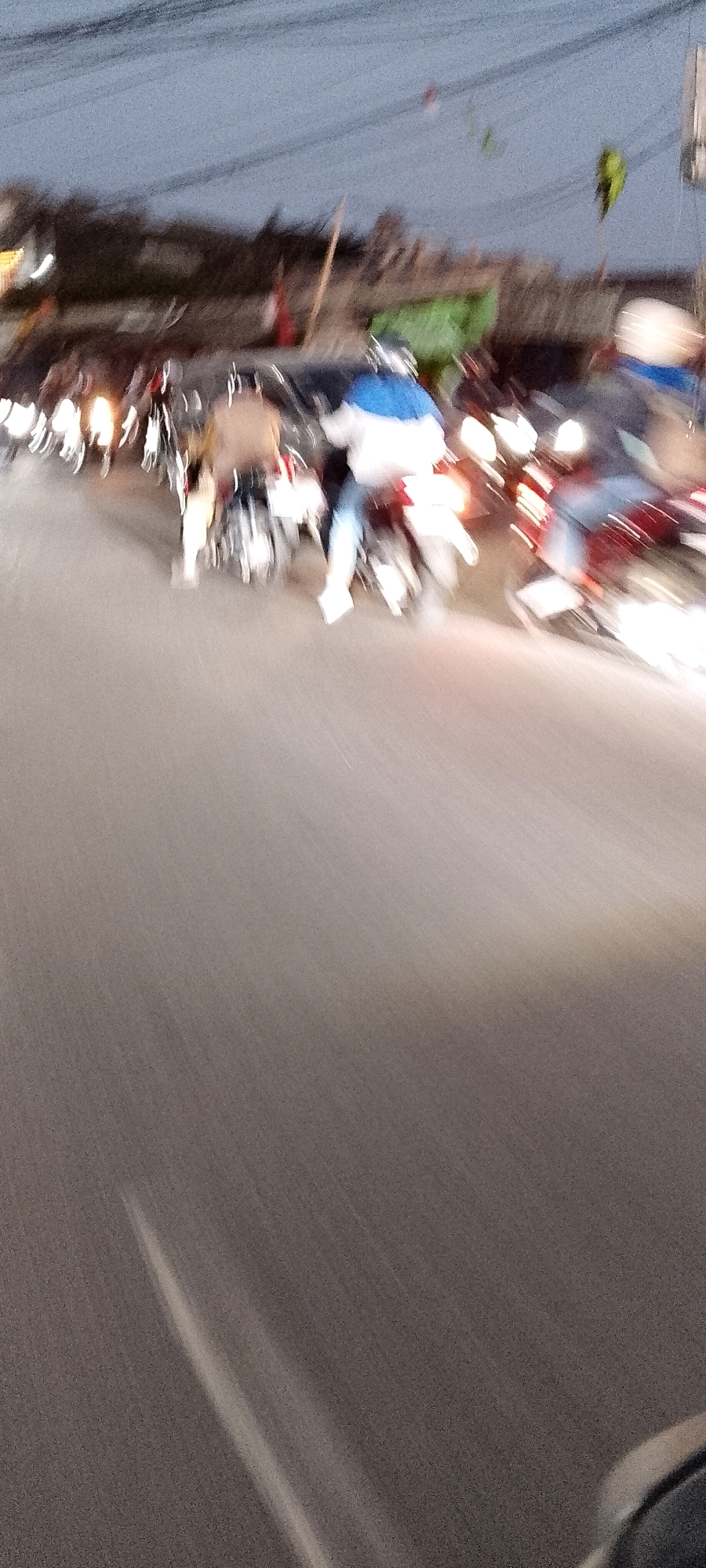 The image depicts a street scene in Depok, Indonesia, characterized by motion blur, suggesting movement of vehicles and pedestrians. Several motorcycles are visible, with riders dressed in casual attire, including a person wearing a blue and white jacket and another in a tan-colored top. The lighting indicates it is either dusk or early evening, with artificial lights from the motorcycles illuminating the road. The background shows a blurred line of buildings and trees, consistent with an urban or suburban environment. The road surface appears to be asphalt. The motion blur is significant across the entire image, making precise identification of individual features difficult, but the overall impression is of traffic on a road.