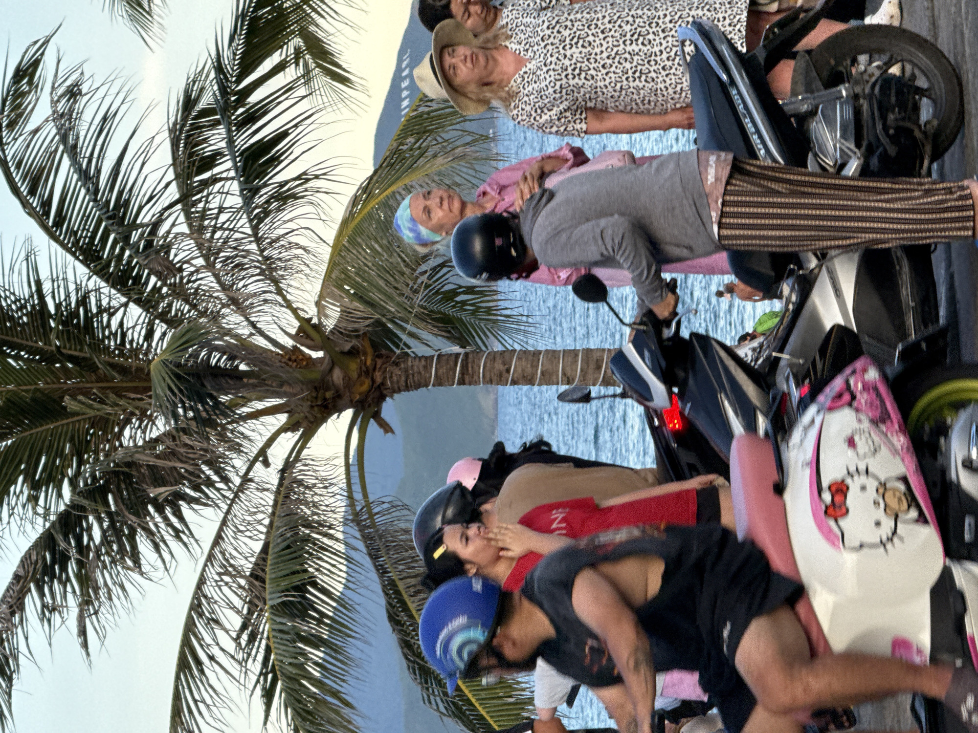 The image depicts a street scene in Nha Trang, Vietnam, likely near the coast. A group of people are gathered near several parked motorbikes. In the foreground, a person wearing a black t-shirt and black shorts is seated on a white and pink motorbike adorned with Hello Kitty graphics. To their right, another individual wearing a grey hoodie and striped pants stands next to a black motorbike. Behind them, a woman in a pink outfit and a patterned headband is looking to the side. Further back, a woman in a leopard print dress and a straw hat is visible. Palm trees with green fronds are prominent, silhouetted against a light blue sky and a body of water, suggesting a coastal setting. In the distance, hills and what appears to be a building with lettering are discernible. The overall atmosphere suggests a casual gathering in a tropical urban environment.