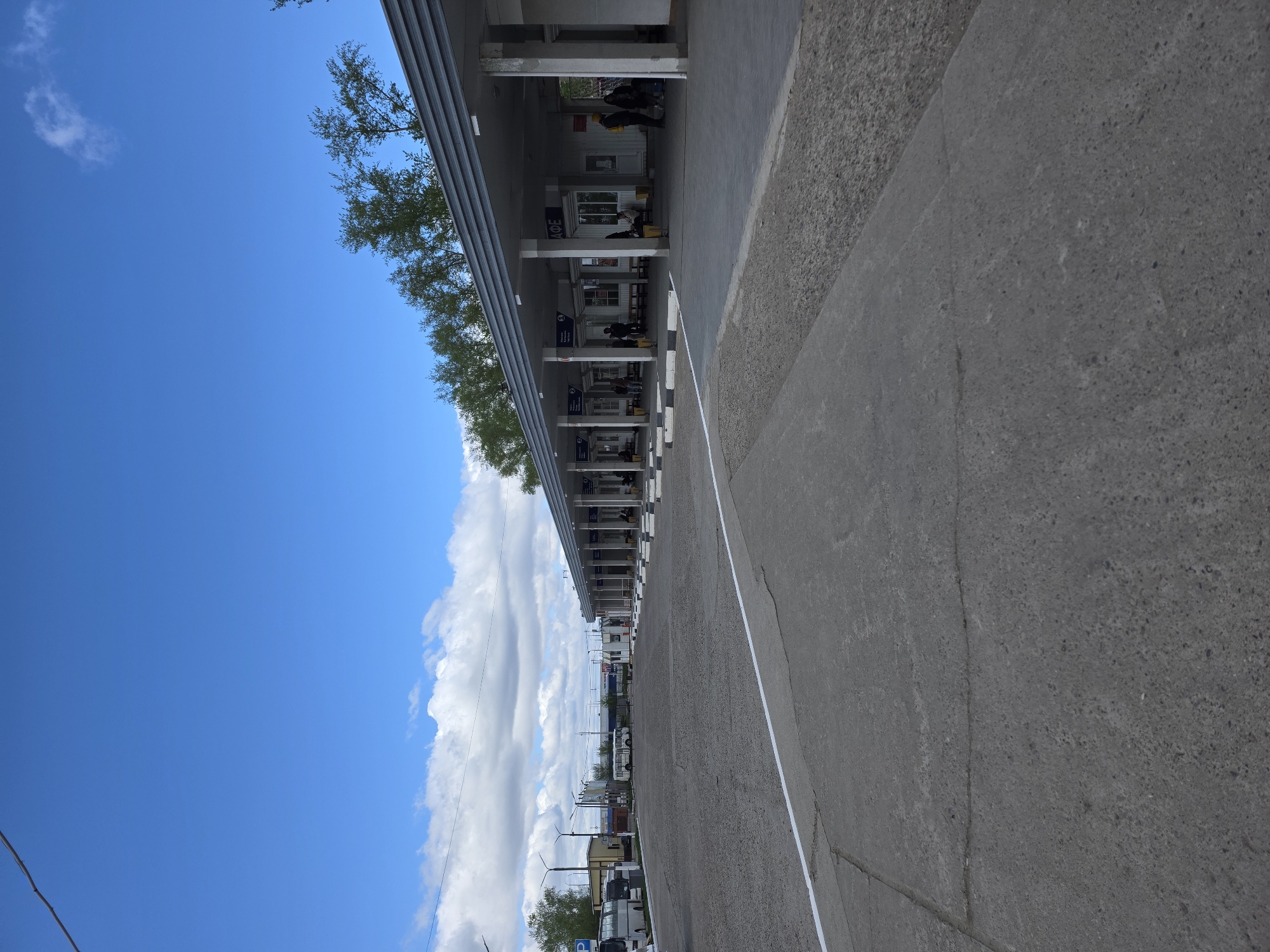 The image shows a long, open-air building with a series of storefronts, likely a market or transportation hub, located in Chaikovsky, Russia. The building has a gray, corrugated metal roof supported by concrete pillars, creating a covered walkway in front of the stores. Signs in Russian script are visible above the entrances. People can be seen indistinctly within the building.
To the right of the building is a wide, gray concrete surface, potentially a road or large walkway, with a white painted line marking its edge. The sky above is partly cloudy with a clear blue expanse. The perspective suggests the photograph was taken at ground level.