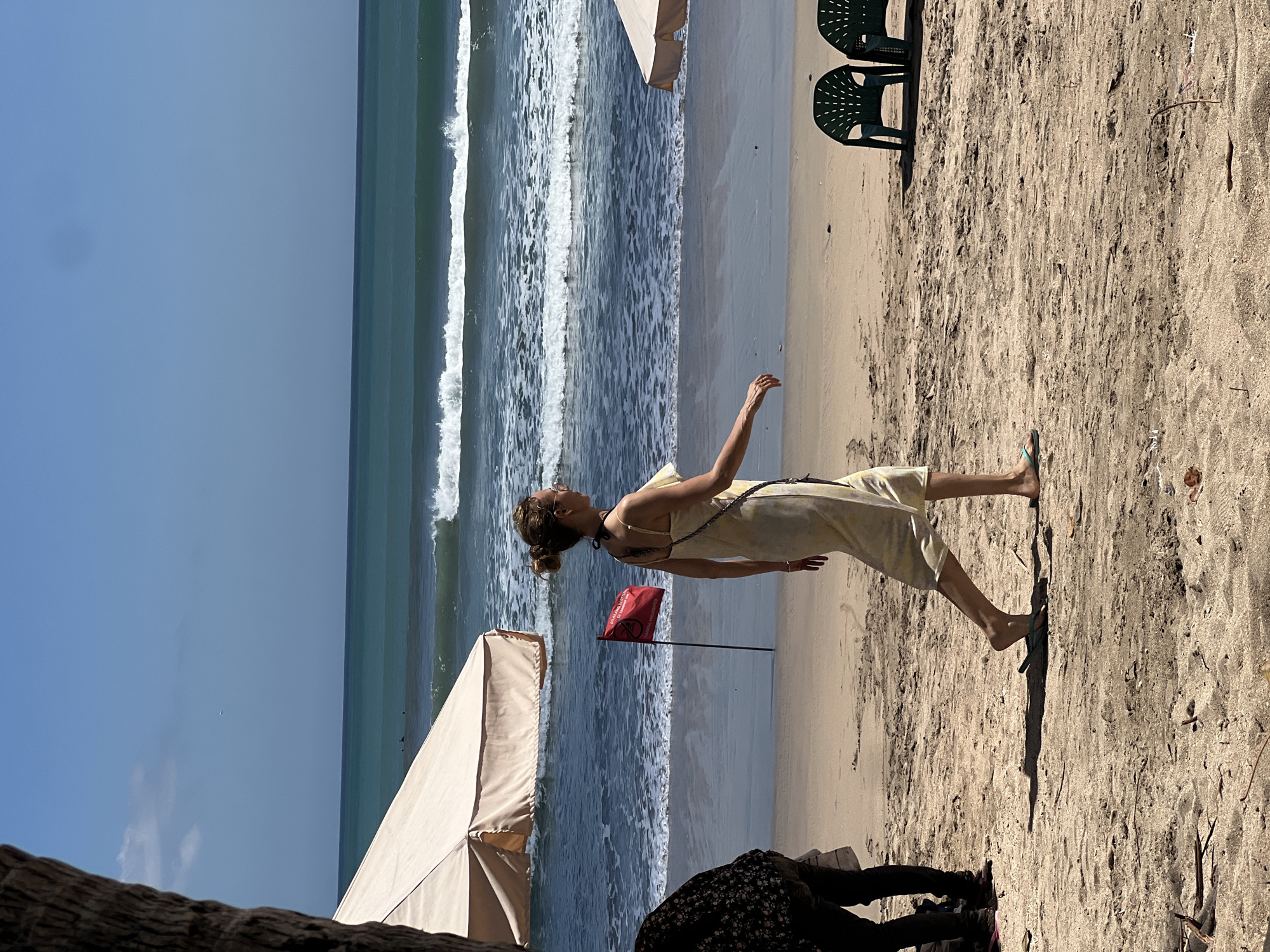 A woman in a light-colored sundress walks along the beach in Kuta, Indonesia, on a bright, sunny day. The ocean behind her features moderate waves. A red flag is planted in the sand, indicating a warning. Two empty green plastic chairs sit on the sand to the right of the woman. In the background, there are two large beach umbrellas. Another person, dressed in dark clothing, is partially visible to the left of the frame. The sand is light brown and appears dry.