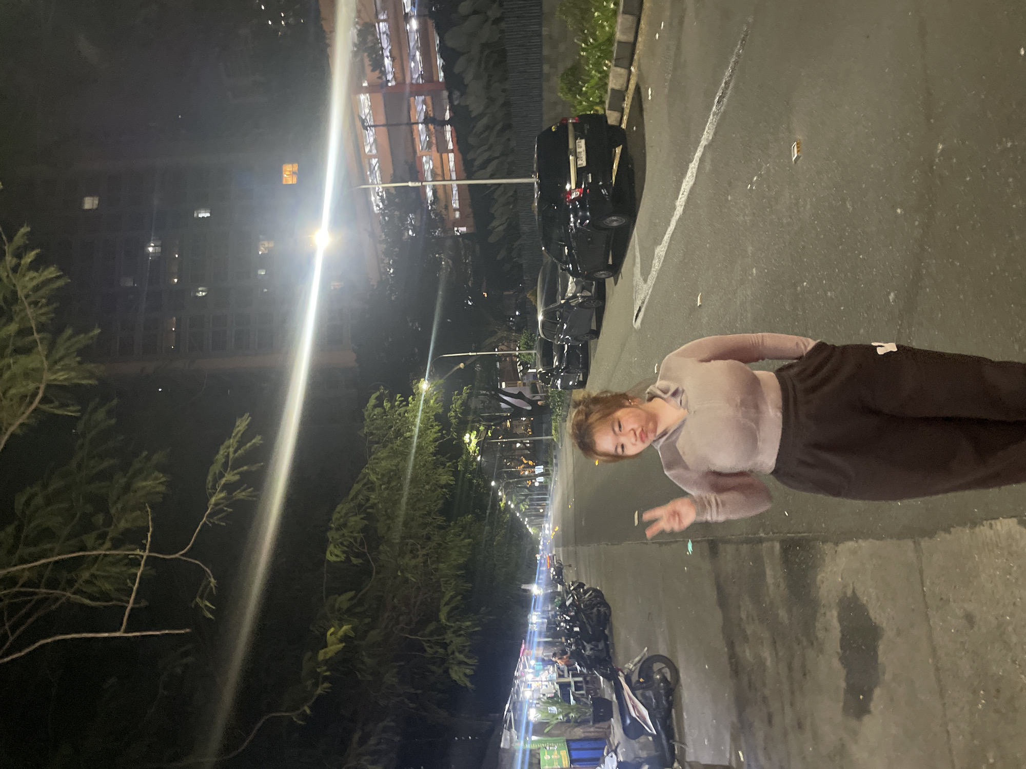 A vertical nighttime photograph shows a young woman posing with a peace sign on a street in North Jakarta, Indonesia. The woman has light skin and wears a light-colored long-sleeved shirt and dark pants. She faces toward the left of the frame, her body oriented horizontally against the dark, wet pavement.
Behind her, a row of parked motorcycles lines the curb, with lights illuminating shopfronts further down the street. Cars are parked along the road beyond the motorcycles. Trees line the street, and tall buildings are visible in the background under the night sky. The overall lighting is dim, punctuated by streetlights and storefront illumination.