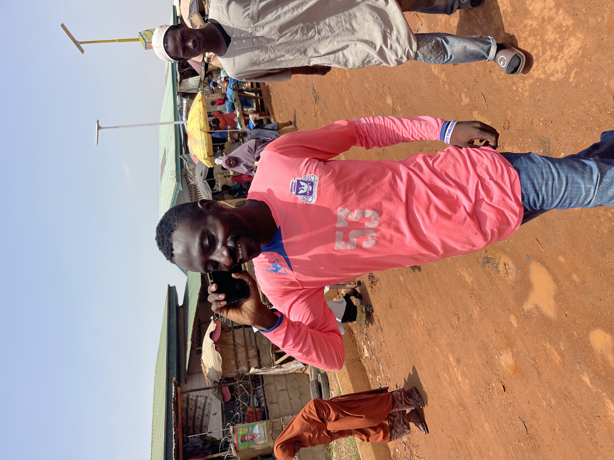 The image depicts an outdoor scene in Zaria, Nigeria. The foreground features a man wearing a pink long-sleeved shirt with blue accents, blue jeans, and a bracelet. He is holding a black smartphone to his ear, appearing to be on a call and smiling. Adjacent to him is another person in a beige outfit, wearing a white cap and jeans.
The backdrop reveals a bustling marketplace environment. Multiple individuals are visible under yellow umbrellas. Structures with green corrugated iron roofs are present. A signpost with a yellow arm extending from the top is visible.
The ground is a mix of red soil, with pools of water. The overall impression is that the photo was taken in a busy, possibly informal, commercial area in Zaria.