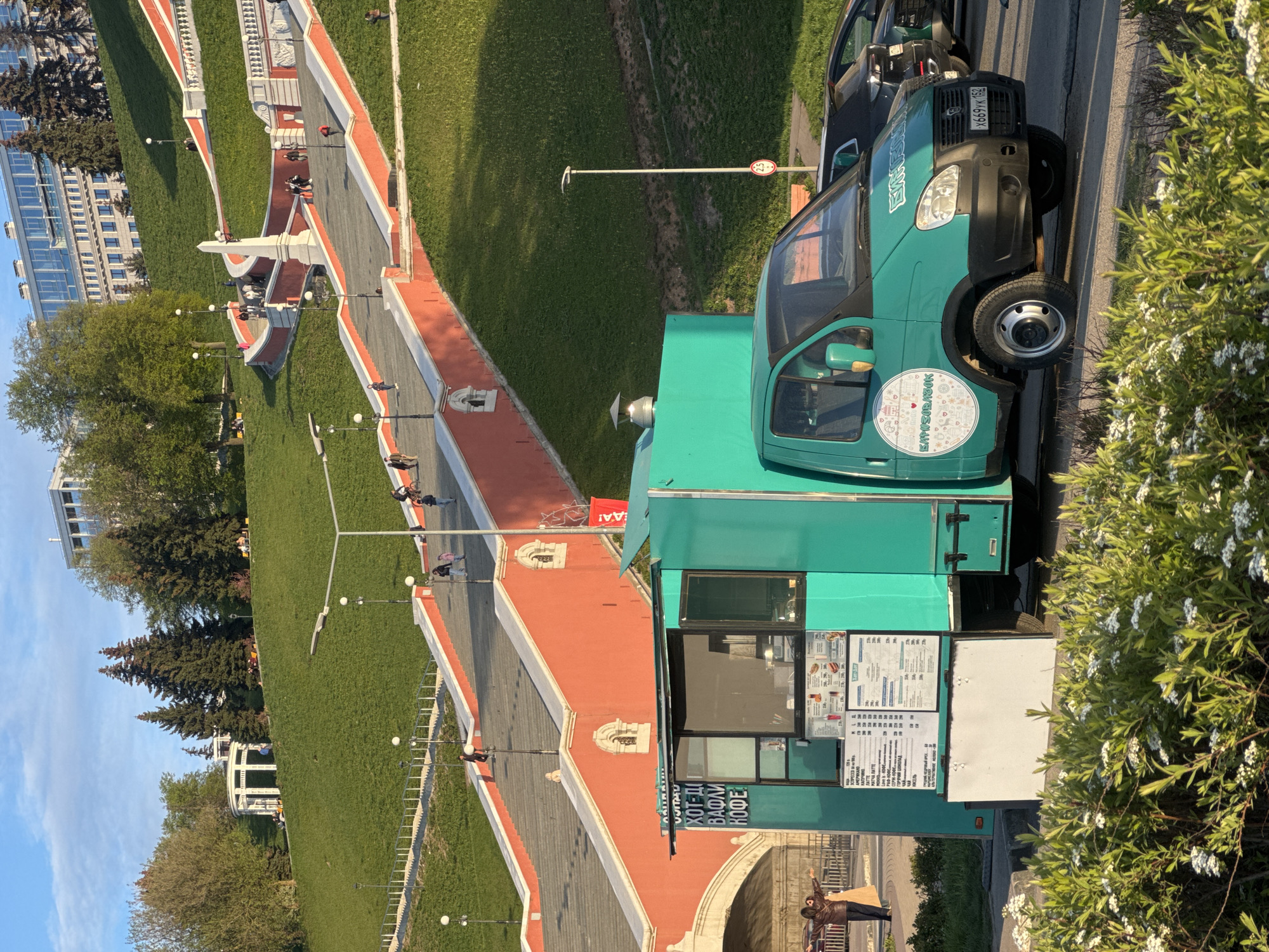 The image shows a turquoise-colored food truck parked on the side of a road with a view of Chkalov Staircase in Nizhny Novgorod, Russia. The truck has a menu displayed with items such as hot dogs, waffles, and coffee. A black car is parked directly behind the truck, and on the license plate you can read: "X669yk 152". The staircase is surrounded by green grass, trees, and buildings. A street sign is visible in the background. People can be seen walking up and down the staircase. The sky is blue with some clouds.
