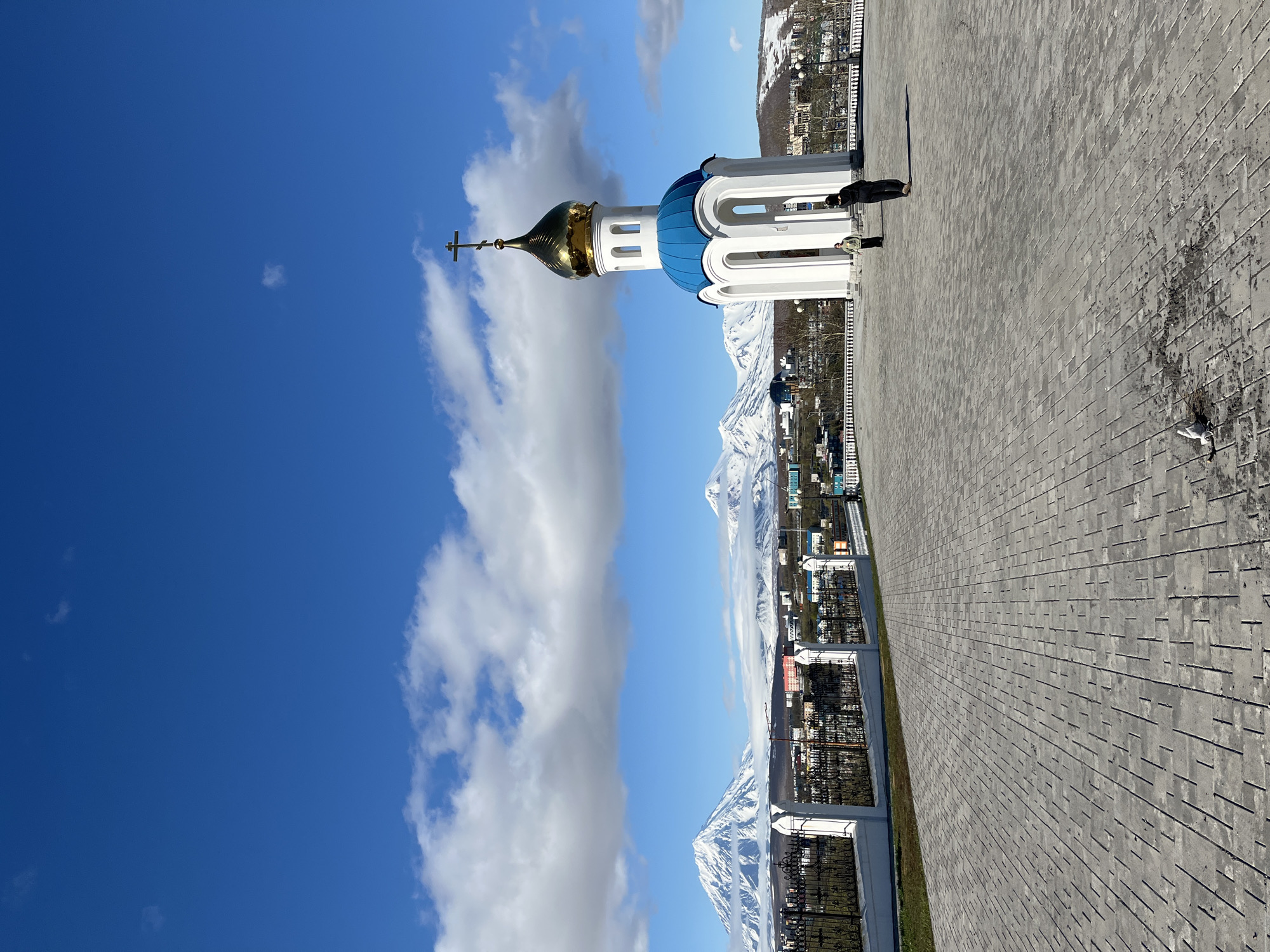 The image shows a white church with a gold dome and blue accents against a backdrop of mountains and a blue sky with white clouds. The location is likely in Petropavlovsk-Kamchatsky, Russia, given the architecture and landscape. Two people are visible, one casting a long shadow on the grey brick walkway. The scene appears to be taken during the day in clear weather. The surrounding environment includes buildings, a fence, and snowy mountain peaks in the distance.