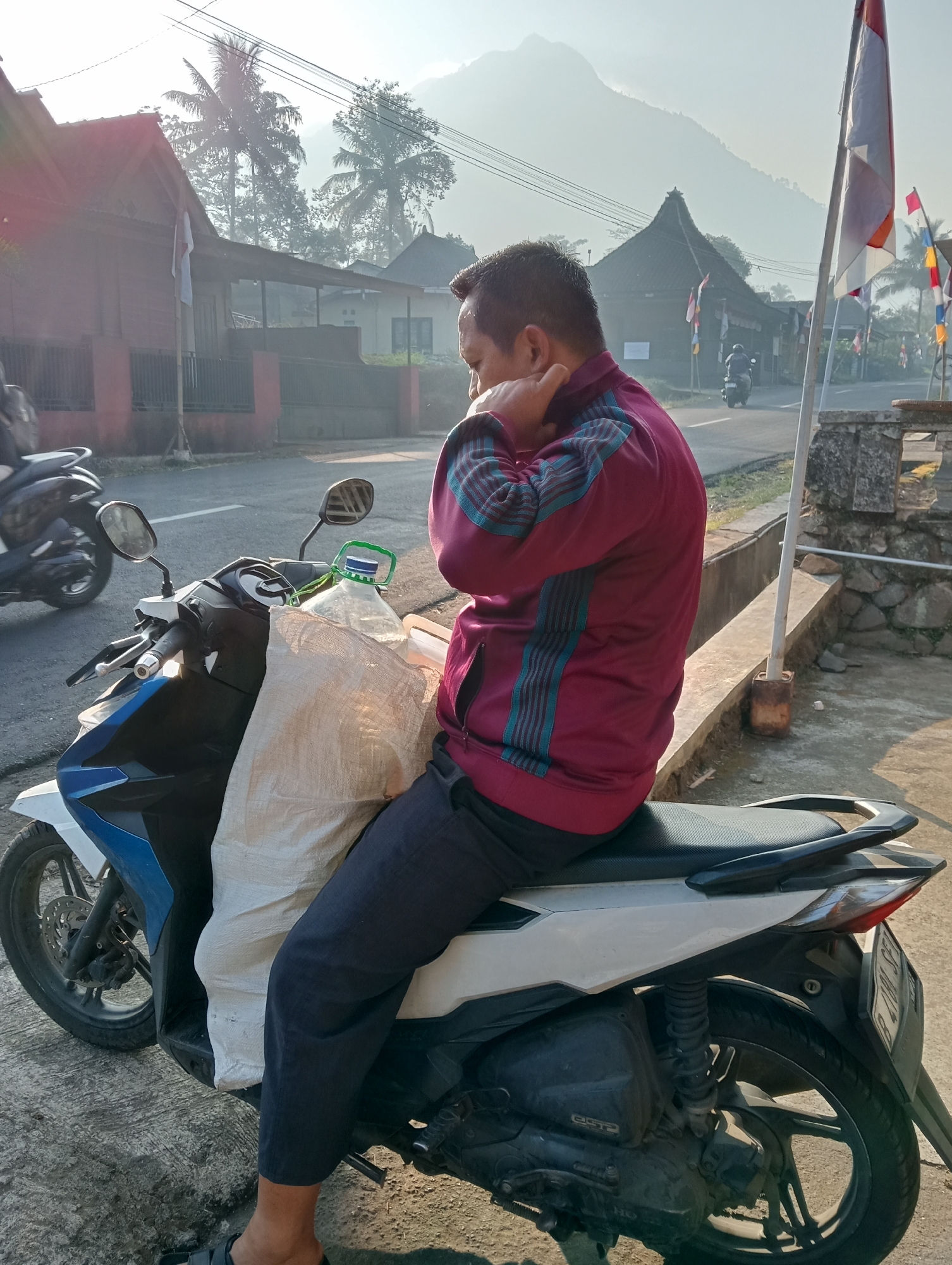 The image depicts a man sitting on a white and blue scooter in the city of Grabag, Indonesia. The man is wearing a maroon jacket with blue stripes and dark pants. He is seen holding his neck with his right hand. The scooter is carrying a large, light-colored sack and a green-handled bottle on its front. In the background, there are traditional Indonesian houses with thatched roofs, palm trees, and a misty mountain range. Several flags are visible, including Indonesian flags. The sun is shining, casting shadows and creating a bright, hazy atmosphere. The scene appears to be a residential street with a few other scooters and motorcycles in the distance.