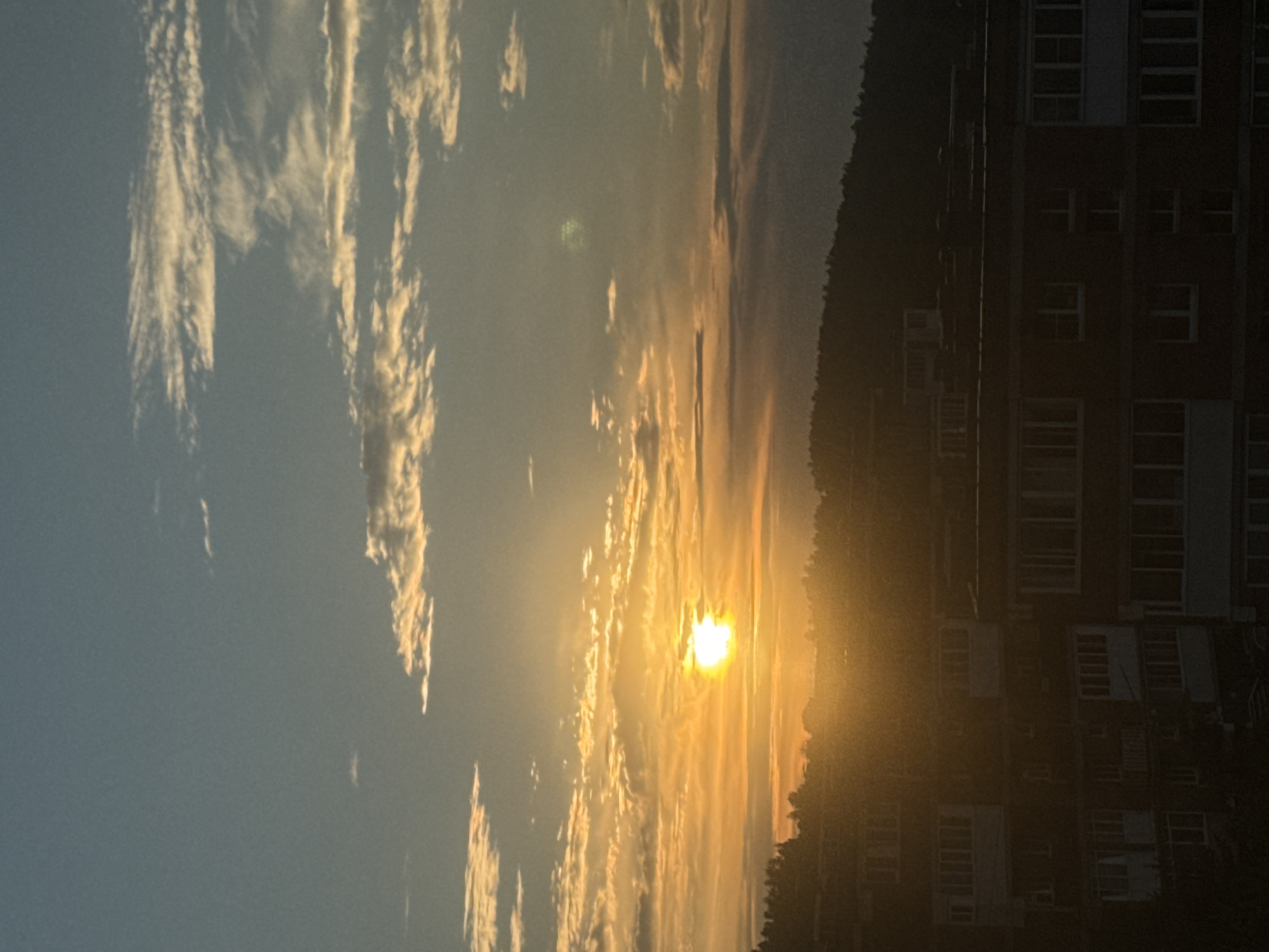 The image depicts a sunset over the city of Markova, Russia. The sun is low on the horizon, casting a warm orange glow across the sky, with scattered clouds tinged with yellow and white. Silhouetted against the vibrant sky is a tree-lined hill or mountain range in the background. In the foreground, the upper floors of several apartment buildings are visible, their dark facades contrasting with the bright sky. The buildings have numerous windows and balconies, suggesting a residential area. The scene is captured from a slightly elevated perspective, looking towards the west.