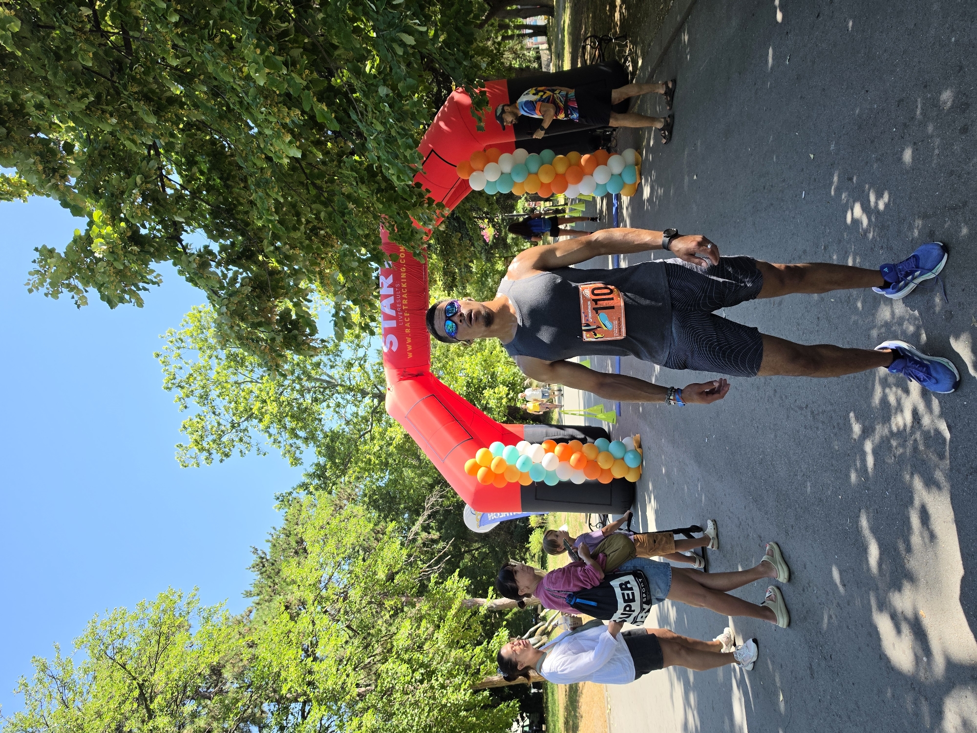 In Varna, Bulgaria, on a sunny day, a male runner with bib number 110 stands at a start line beneath a large red inflatable arch. The arch is decorated with balloons in orange, white, and teal. The runner wears a grey tank top, patterned shorts, sunglasses, and blue athletic shoes. He has a watch on his left wrist and a ring on his left ring finger. To his left, two women and a child stand. The woman closest to the camera wears a white shirt and dark shorts, while the woman next to her wears a pink top, denim shorts, and carries a black bag. The child is dressed in brown shorts and a purple shirt. Behind the runner, another person in a colorful shirt and black shorts stands near the arch. Trees with green foliage line the background, and the scene takes place on an asphalt road, possibly within a park.