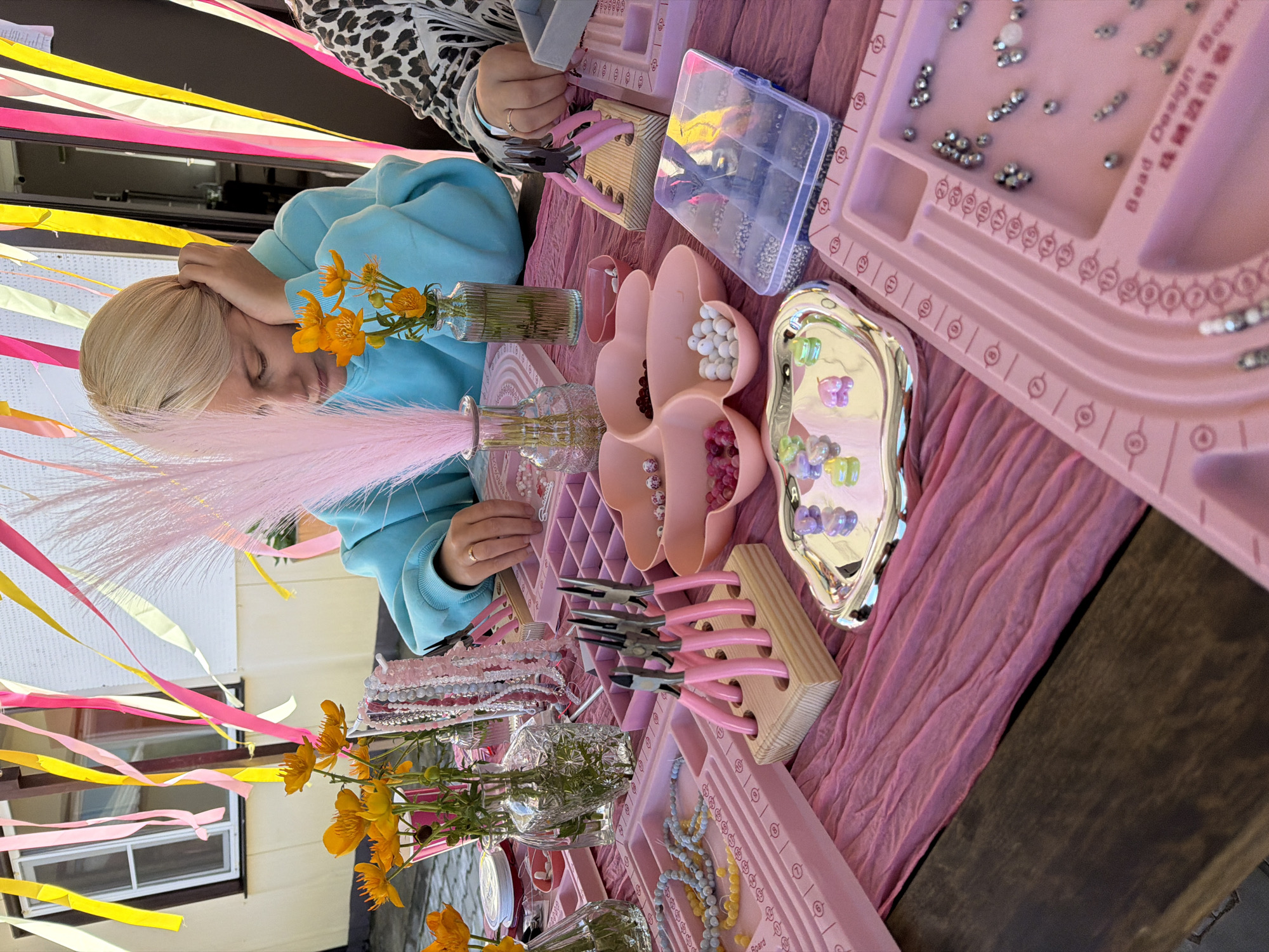 In Birobidzhan, Russia, a blonde woman in a blue hoodie sits at a table covered with a pink textured cloth, engaged in beading or jewelry making. She rests her head on her hand, seemingly deep in concentration. The table is adorned with various crafting supplies: pink plastic trays with compartments for beads of different colors and shapes, jars of beads, a silver tray filled with translucent heart-shaped beads, a wooden stand holding pink-handled jewelry pliers, and several vases with yellow flowers. Decorative streamers in yellow, pink, and cream hang overhead, suggesting a festive or creative gathering. Another person, partially visible on the right, is also participating in the activity, their hands and a leopard-print sleeve visible. The scene takes place outdoors, with a building and a window visible in the background.