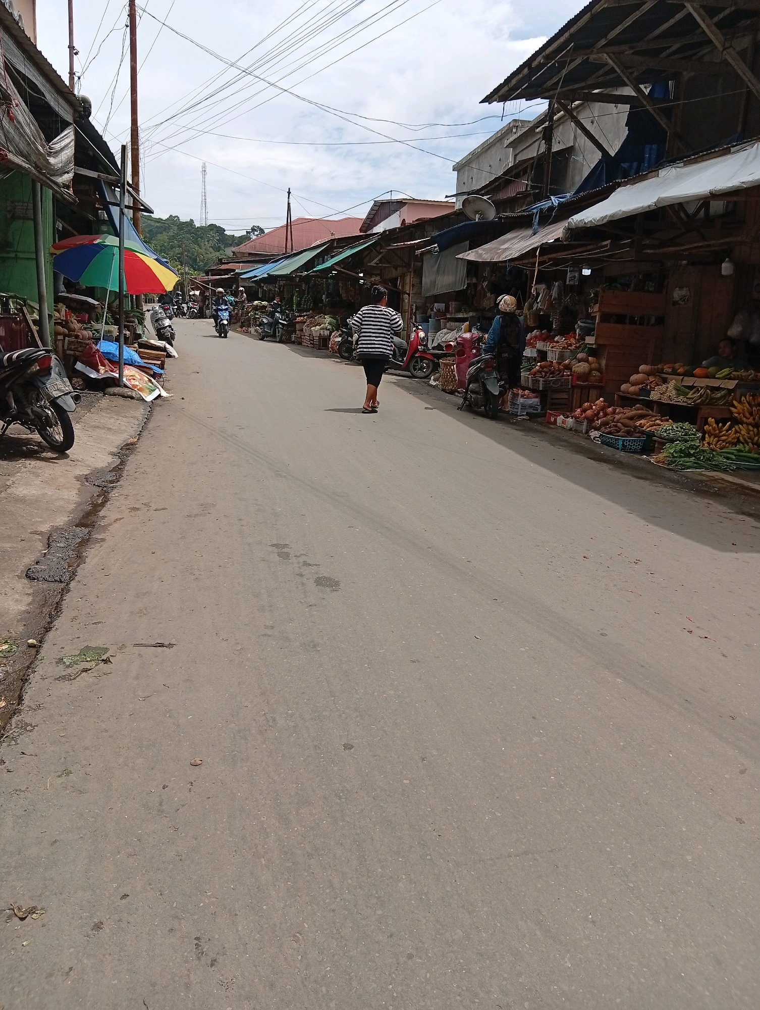 The image shows a street scene in the City of Gunungsitoli, Indonesia, likely a market area. The street is paved and appears to be two lanes wide. On either side of the street, there are stalls or shops with open fronts displaying various produce, including fruits and vegetables. Several motorcycles are parked along the sides of the road, and some are in motion on the street. People are seen walking and interacting with the market stalls. The architecture of the buildings suggests a tropical or Southeast Asian setting, with wooden structures and corrugated metal roofs. Power lines are visible overhead. The sky is overcast. One person in the foreground is walking away from the camera, wearing a striped shirt and dark pants. Another person is seen on a scooter near the market stalls. The overall impression is of a bustling, everyday market scene in a town.