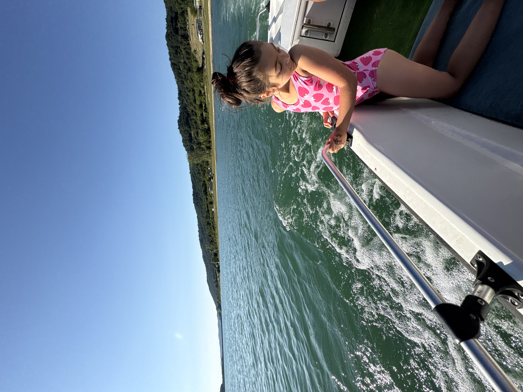 A young girl with dark hair in a ponytail, wearing a pink swimsuit with a strawberry pattern, leans over the edge of a white boat. She is looking down at the water, her eyes closed as if enjoying the sensation of the boat moving. The boat is on a body of water, possibly a lake or river, with green, tree-covered hills in the background. The sky is clear and blue, indicating a sunny day. The water is choppy, with white foam and ripples created by the boat's movement. A metal railing runs along the side of the boat, and the girl's hand is resting on it. There is a tattoo of a mermaid on her hand. The scene suggests a recreational activity, likely a boat ride on a warm day, possibly in the summer. The overall mood is peaceful and relaxed.