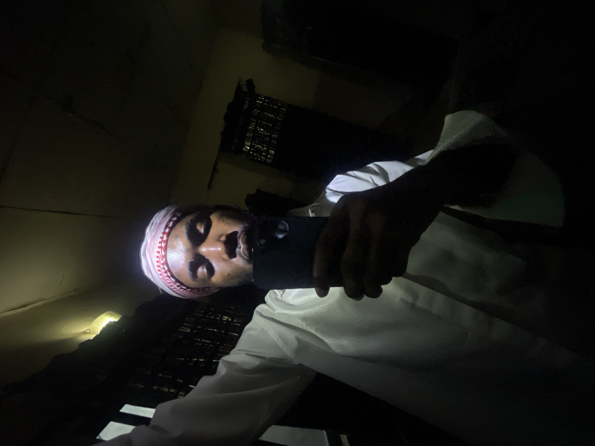A person wearing a red and white head covering is holding a black smartphone and taking a selfie in a dark room. The room has a dark window with blinds and a light is shining on the wall. The location is Unknown, Unknown.