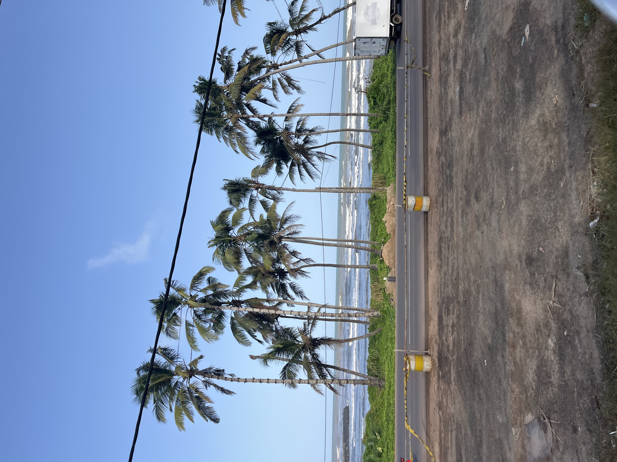 The image depicts a coastal scene in Maggona, Sri Lanka, with a row of palm trees lining a road that runs parallel to the ocean. The sky is clear and blue, with a few wisps of clouds. The ocean is calm with gentle waves breaking on the shore. A road is visible in the foreground, with yellow and black caution tape stretched across it, supported by yellow and white barrels. A white truck is parked on the right side of the road. The ground in the immediate foreground is uneven and appears to be dirt or gravel, with some sparse vegetation. Overhead power lines run across the top of the frame. The overall impression is one of a tropical, seaside location.