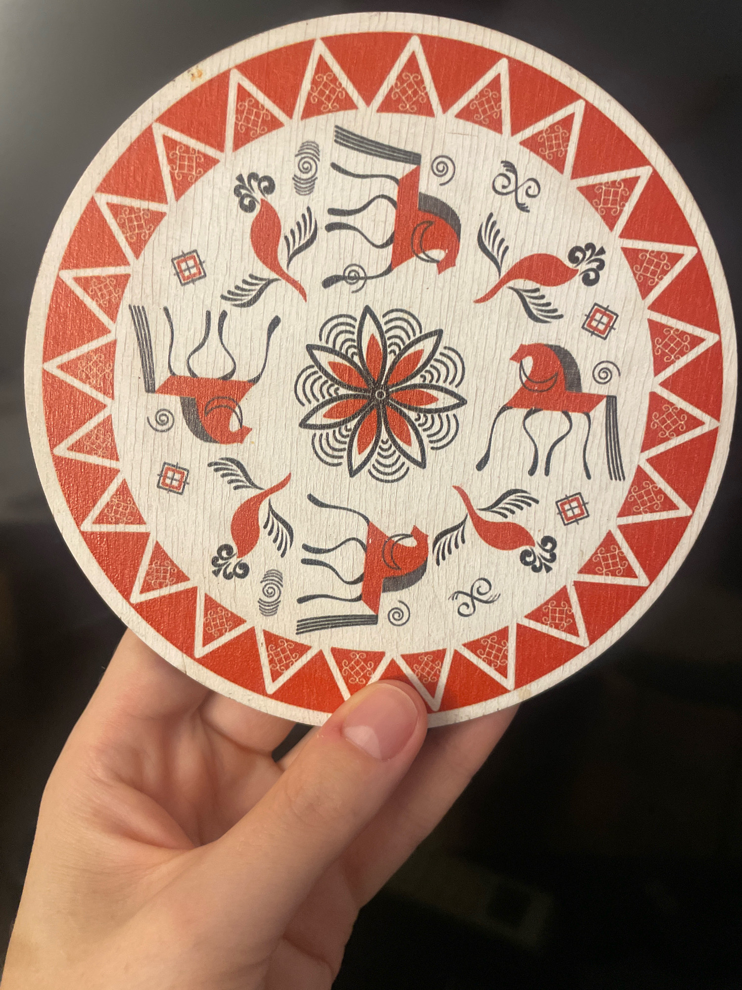 A hand is holding a circular coaster with a red and white folk art design. The design features stylized red birds and horses, along with black geometric patterns and a central floral motif. The coaster is made of wood and has a light-colored grain. The background is dark and out of focus. The image does not contain any information that would indicate its location in Atarfe, Spain.