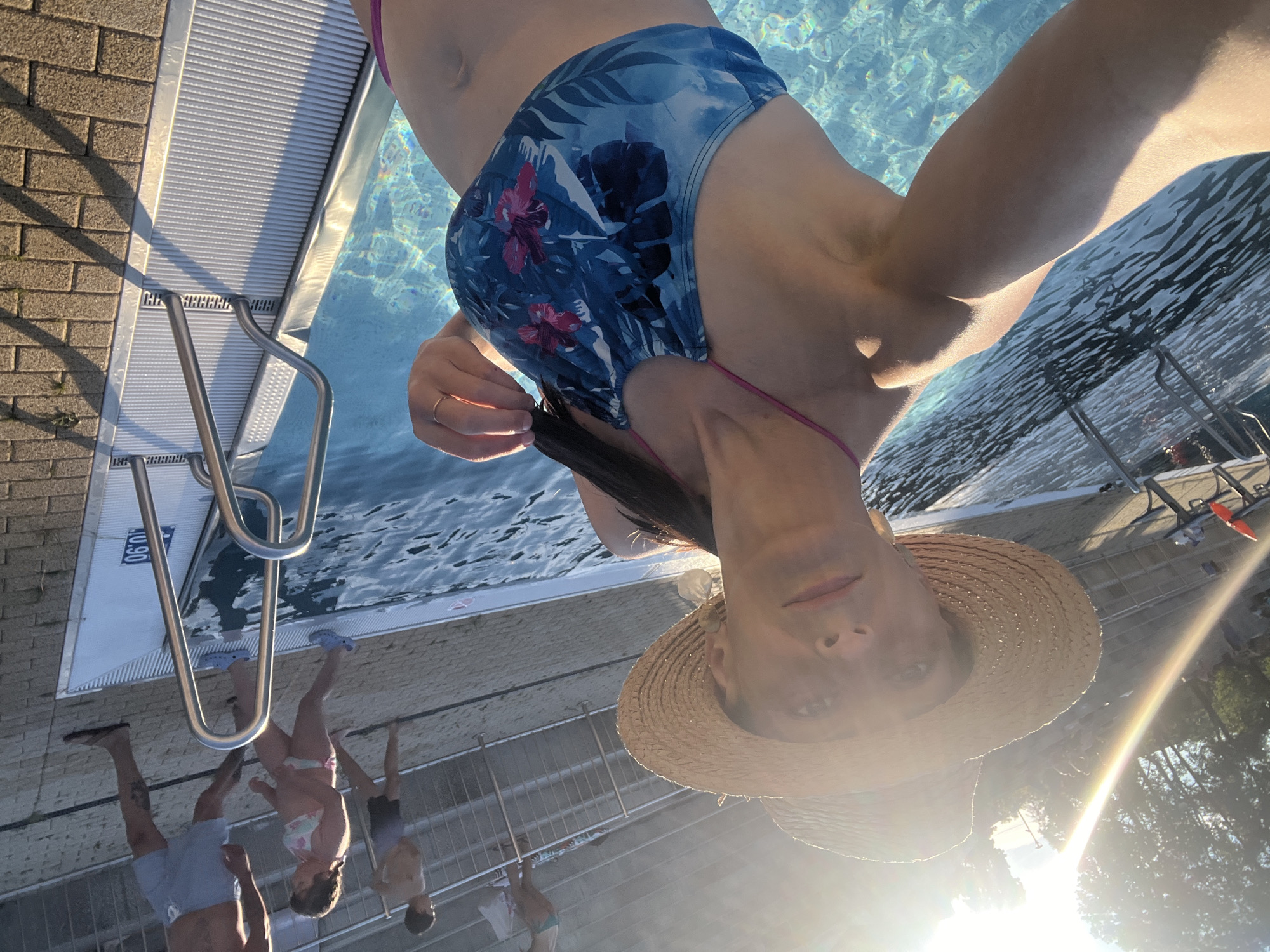 A woman wearing a straw hat and a blue floral bikini top poses for a selfie by a swimming pool. She is partially submerged in the water, and her right hand is raised, touching her hair. The sun is shining brightly, casting long shadows. In the background, several people are visible near the pool steps and on a higher level. The pool area has brick paving and metal railings. The image is taken from a low angle, looking up at the woman.
