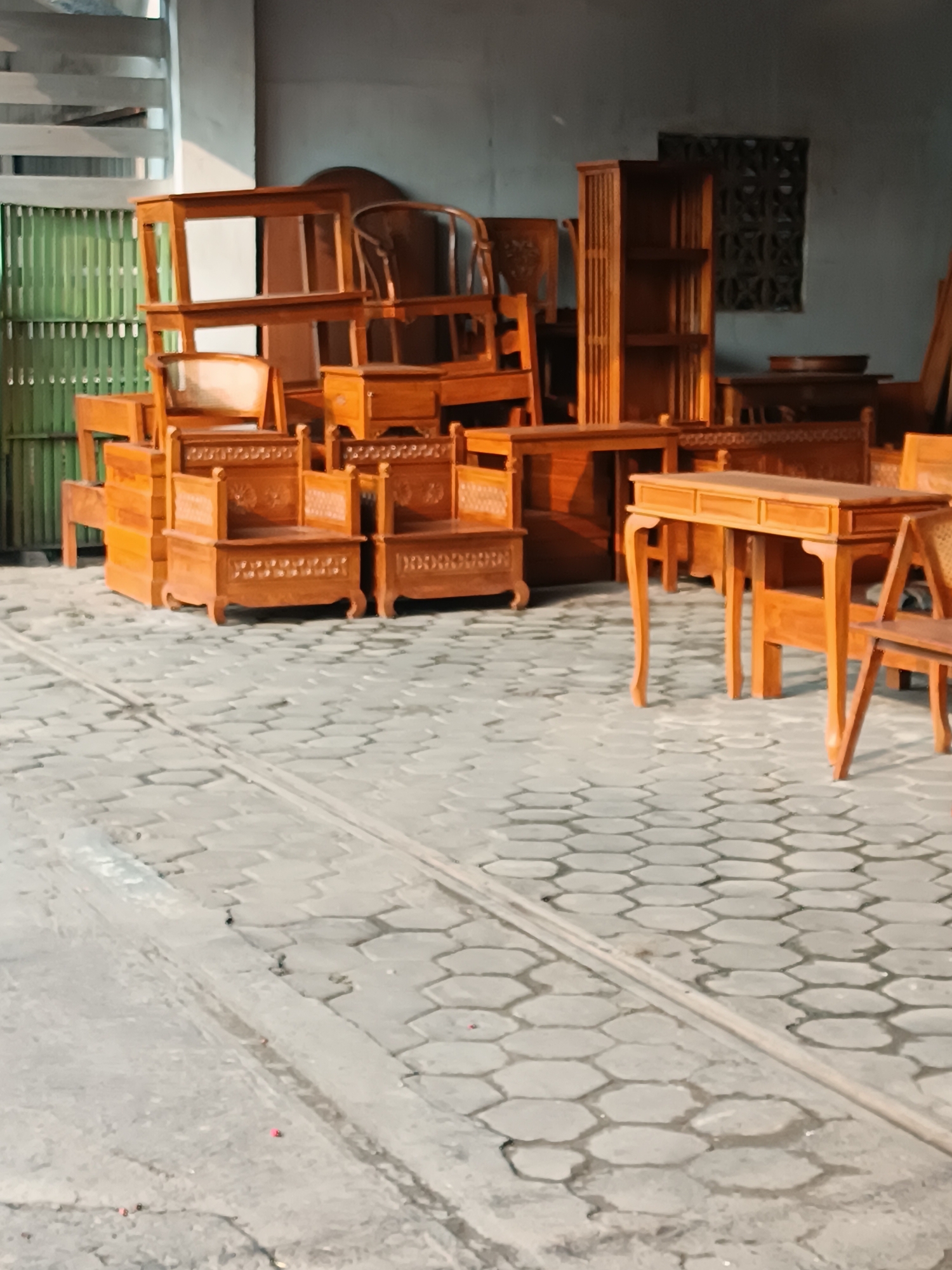 In Banguntapan, Indonesia, a collection of wooden furniture is displayed outdoors on a paved area. The furniture, predominantly made of light-colored wood, includes chairs, tables, and shelving units. Two intricately carved chairs with backs are prominent in the foreground. Behind them, a stack of wooden boxes or drawers is visible. To the right, a writing desk with curved legs stands alongside another wooden chair. In the background, a tall, slatted bookshelf is positioned against a plain wall, with a decorative grilled window above it. The ground is covered with hexagonal paving stones, and a light-colored vertical structure, possibly a gate or screen, is on the left side of the image. The scene appears to be a furniture display or storage area.