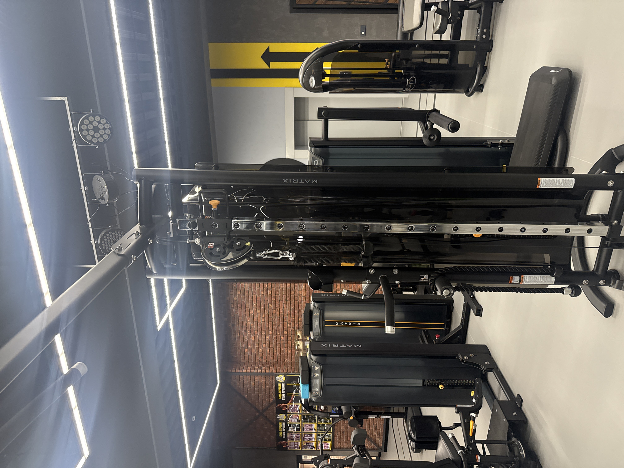 This image shows a piece of "Matrix" brand gym equipment inside a gym. The equipment has an adjustable bench and various bars and pulleys. The gym's interior includes LED lighting on the ceiling, a yellow arrow sign on the wall, and a brick wall section. Posters are also visible behind the gym equipment. The OCR reveals the phrases "БУДЬ ПЕРВЫМ! СТАНЬ ЛУЧШИМ!" (Be the first! Become the best!) in Russian. It's difficult to confirm the city and country. But OCR information indicates the location is Votkinsk, Russia.
