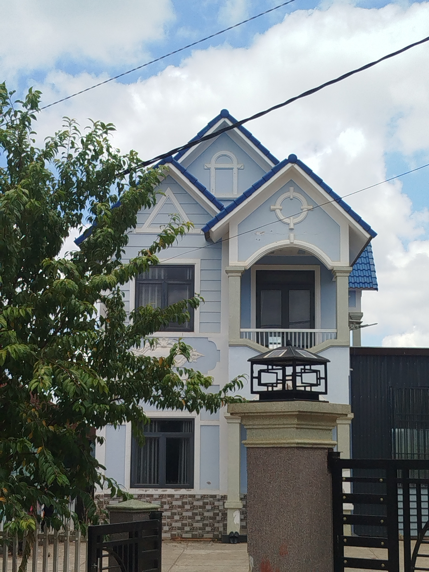 A light blue, two-story house with blue tiled roofs is viewed from the exterior. The house has white trim and grey windows. A large green tree with dense foliage is positioned in the foreground to the left of the house, obscuring a portion of the lower left side of the structure. A black metal fence runs along the bottom of the image. In front of the house, there is a stone pillar topped with a black metal lantern. The sky above is blue with scattered white clouds and a black wire stretches across the upper portion of the frame. The scene is likely a residential area in Thôn 6, Vietnam, given the architectural style and surrounding elements. There are no people visible in the image.