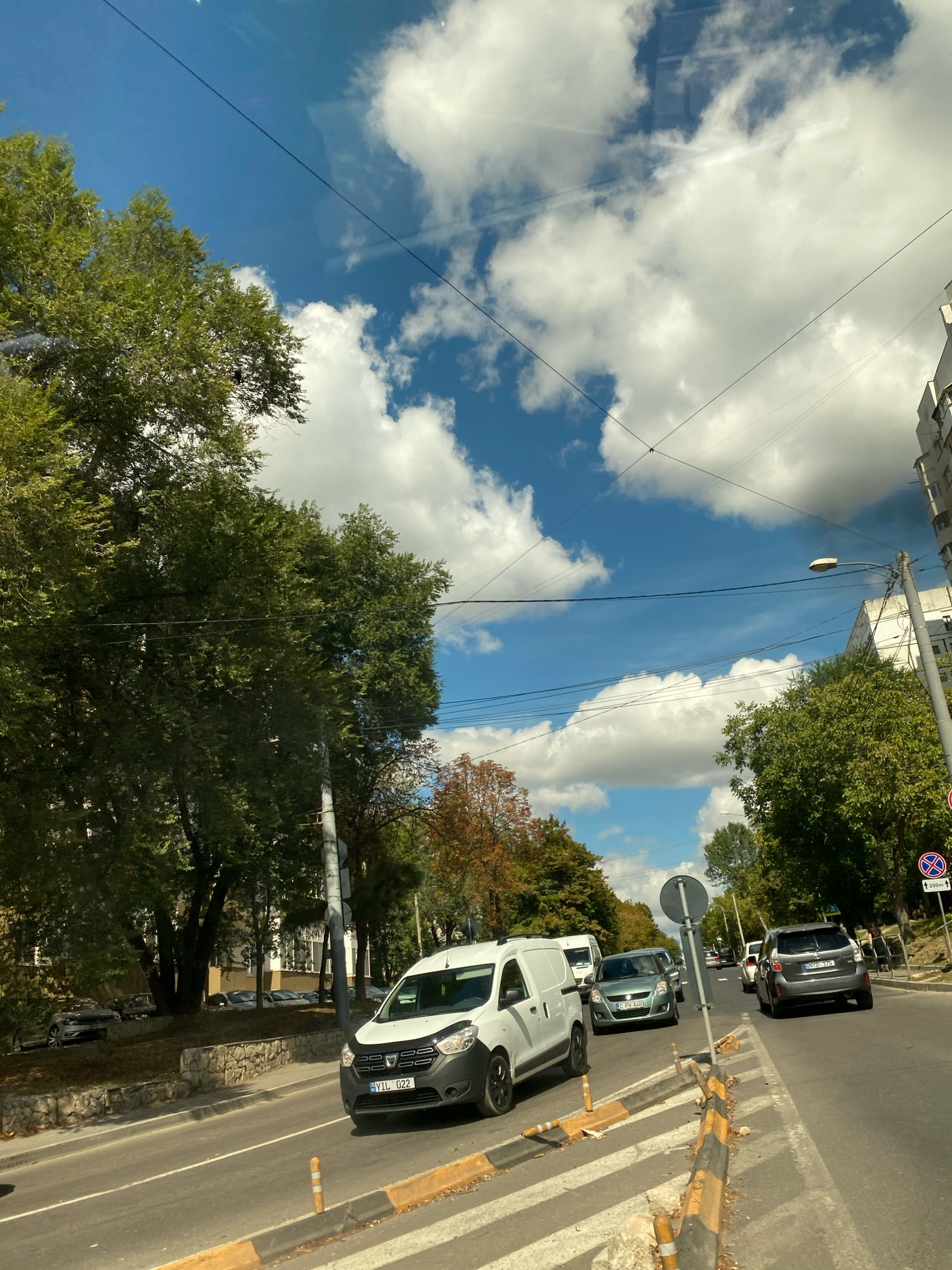 The image captures a street scene in Chișinău, Moldova, on a bright, cloudy day. The perspective is from a vehicle, suggesting movement along a road. In the foreground, a white van with the license plate "VIL 022" is prominently displayed. Beside it, traffic cones and a pedestrian crossing with yellow and black stripes are visible, indicating a road intersection or a construction zone.
The road itself appears to be a multi-lane avenue, with several other vehicles present. To the right of the white van, a grey hatchback car with the license plate "HYD 175" is driving away from the camera. Further down the road, a light green car with the license plate "CPV 640" is also visible. The presence of multiple cars in the same lane suggests moderate traffic.
Lush green trees line the sides of the road, with some autumn foliage starting to appear on some trees, suggesting the season might be early autumn. In the background, a multi-story building with a light-colored facade stands on the right side of the street. Power lines and streetlights are also visible, typical of an urban environment. The sky is a vibrant blue, dotted with fluffy white clouds, adding to the overall bright atmosphere of the scene. The overall impression is that of a typical day in a moderately busy urban setting.