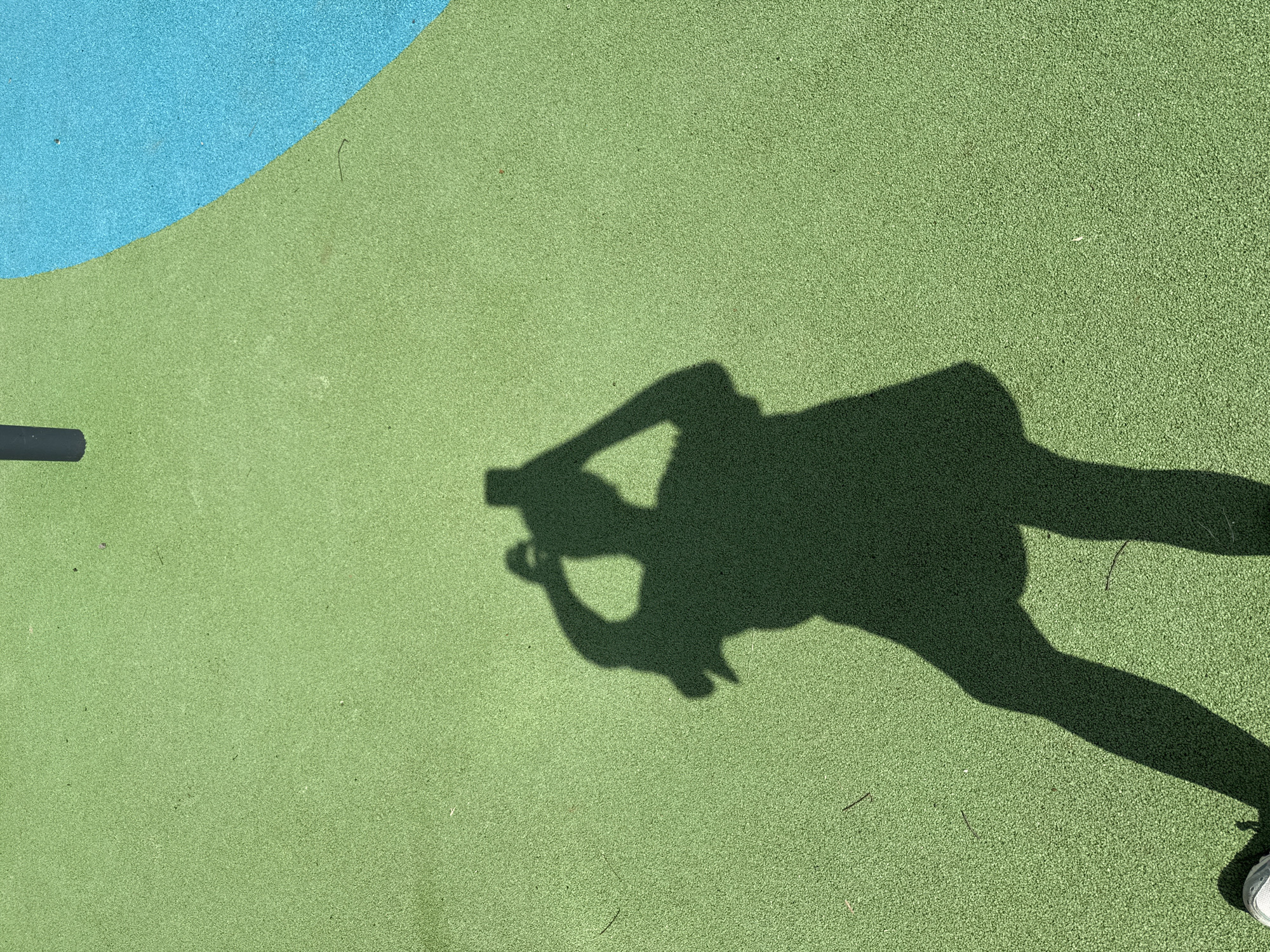 The image shows an overhead view of a playground surface featuring green and blue areas. A person's shadow is cast on the green section. The shadow appears to be holding a phone, and the body is angled towards the camera. A pole is visible on the left side of the image. The location is not possible to identify as Moscow, Russia without additional context.
