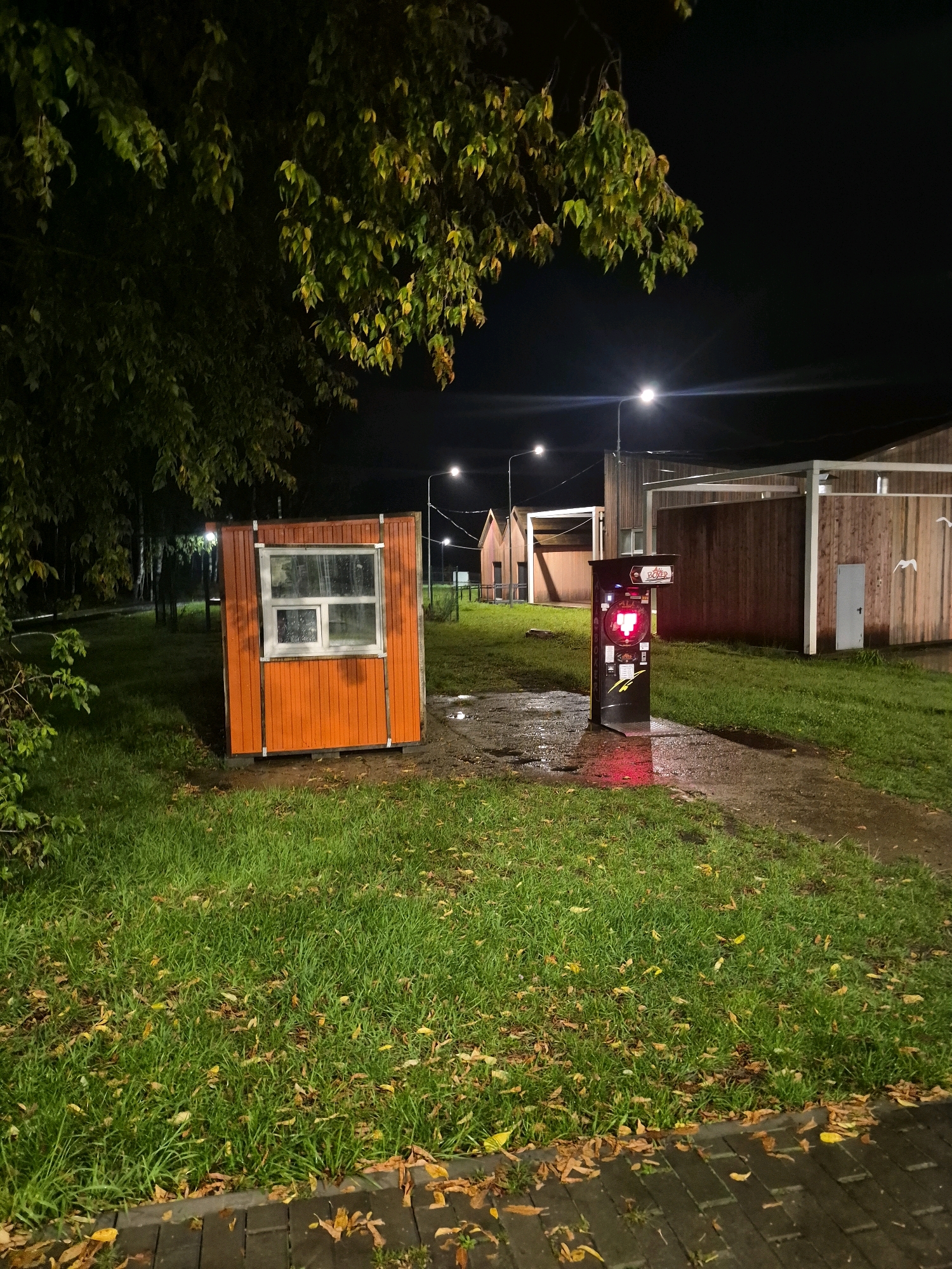 In the city of Chaikovsky, Russia, at night, an orange kiosk with a window sits on a patch of green grass. To its right, a dark, tall machine with a red illuminated display stands on a wet, paved path. Behind these objects, wooden buildings with white awnings line the background. Streetlights illuminate the scene, and trees with green and yellow leaves hang over the left side of the frame. Fallen leaves are scattered across the grass and the pavement.